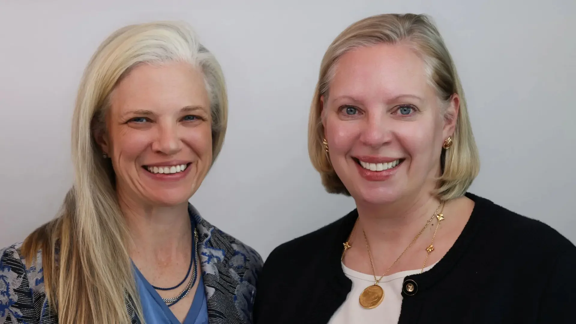 Two women smiling, one with long blonde hair and the other with short blonde hair, standing against a plain light gray background.