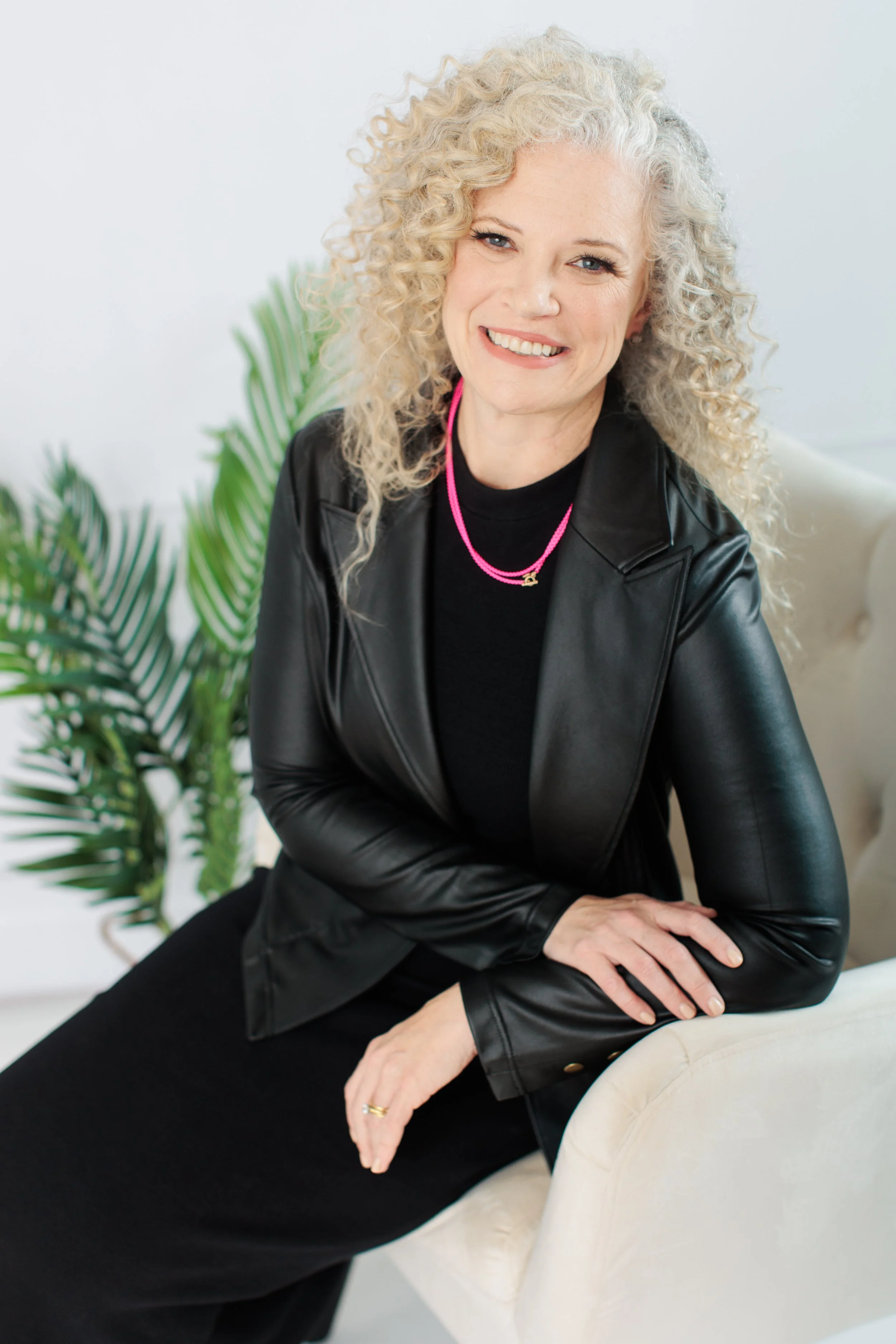 A woman with curly gray hair smiling, sitting on a cream-colored chair, wearing a black leather jacket and a pink necklace, with a green plant in the background.