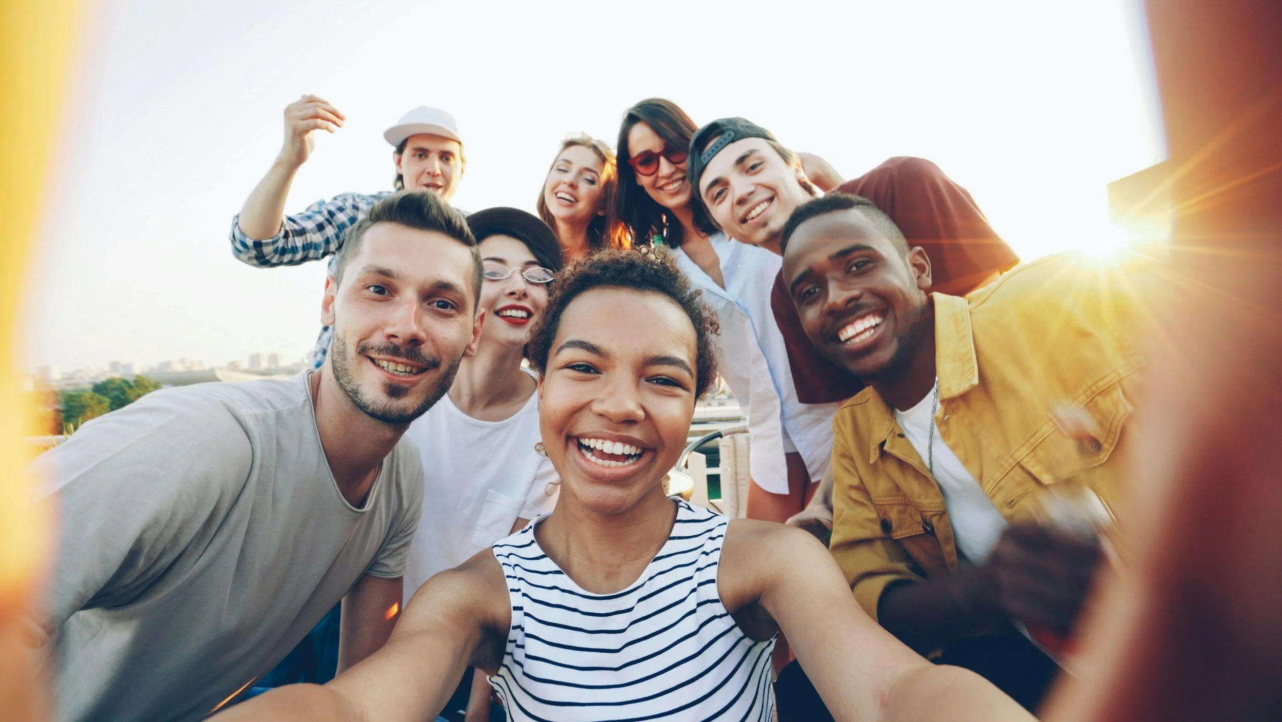 Group of diverse young adults taking a selfie outdoors during sunset, smiling, with buildings in the distance.