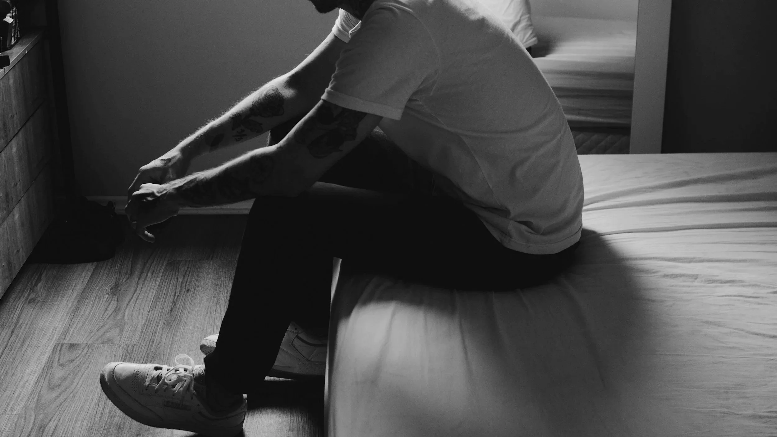 A person with tattooed arms wearing a white t-shirt and white sneakers sitting on a bed, with their head bowed and hands on their knees, in a room with hardwood flooring.