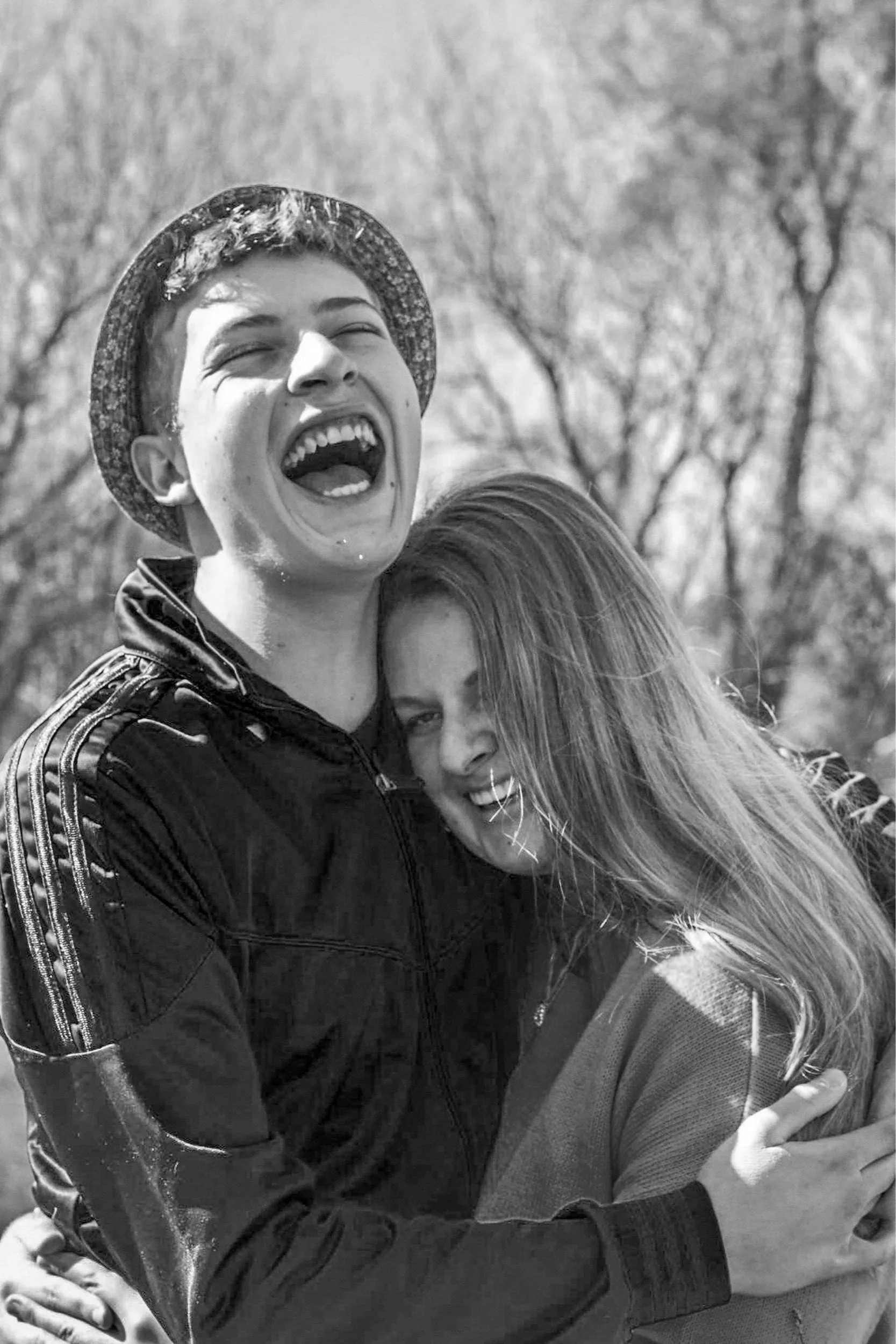 Two young women hugging and smiling outdoors, one with short hair and the other with long hair, with trees in the background.