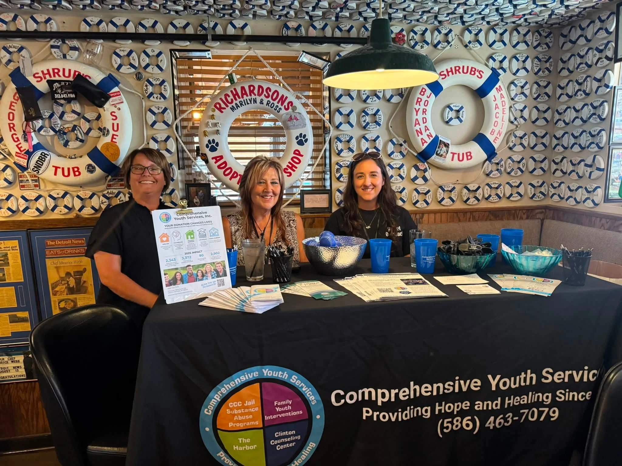 Three women sitting behind a table at an outreach event for youth services, with a decorated wall featuring life preservers with names and messages behind them.