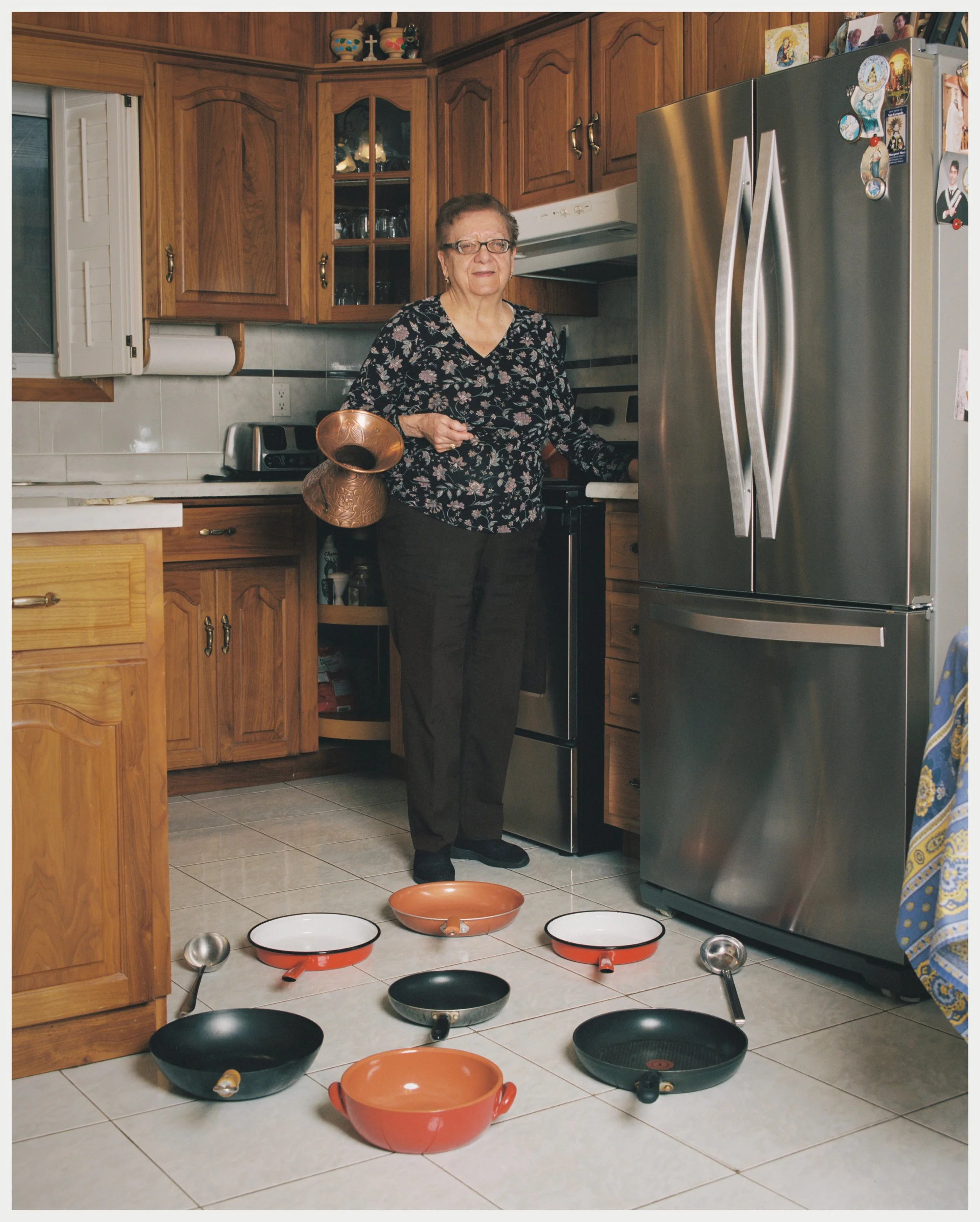 An elderly woman with glasses standing in a kitchen surrounded by fallen cookware, including pots, pans, and ladles, with a sad or disappointed facial expression.