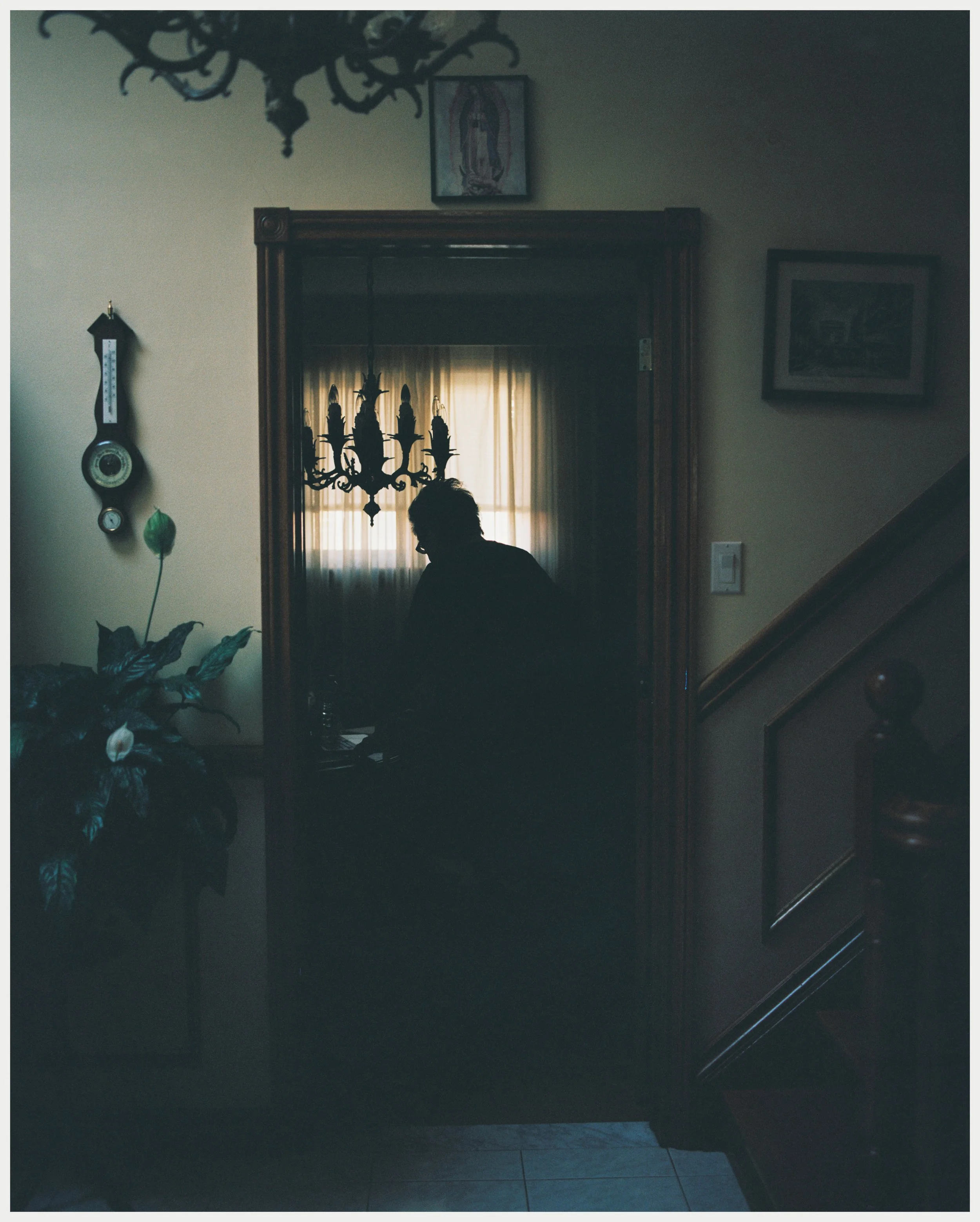 A person sitting at a table in a dimly lit room, with a chandelier hanging overhead. There are pictures and a thermostat on the wall, with curtains covering a window in the background.