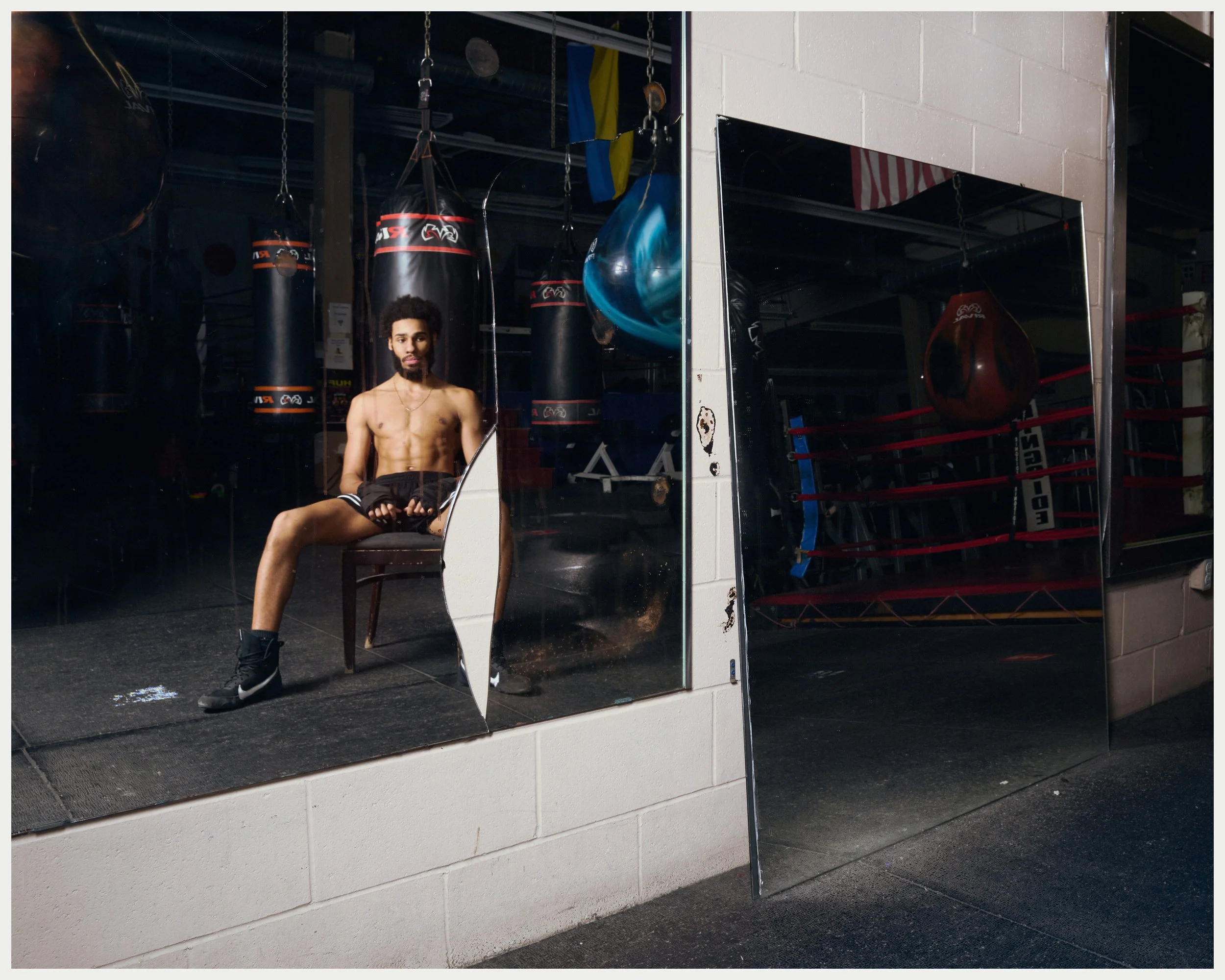 A shirtless man with curly hair and a beard sitting on a chair in front of a gym mirror, with boxing punching bags and gym equipment visible in the background.