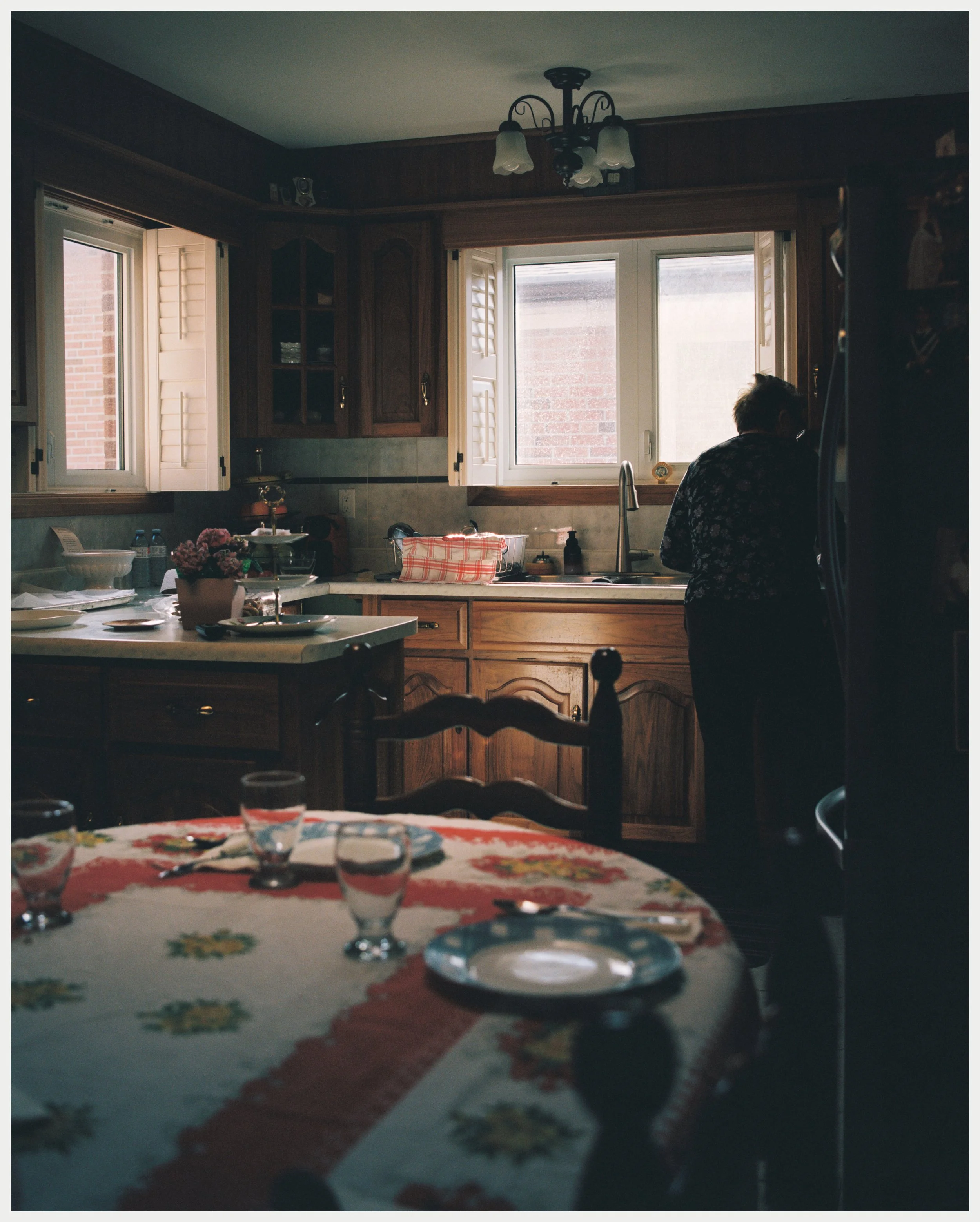 A cozy kitchen with wooden cabinets, two open windows letting in natural light, and a woman washing dishes with a dish towel nearby. The foreground shows a table with glasses, a bowl of flowers, and a floral tablecloth.