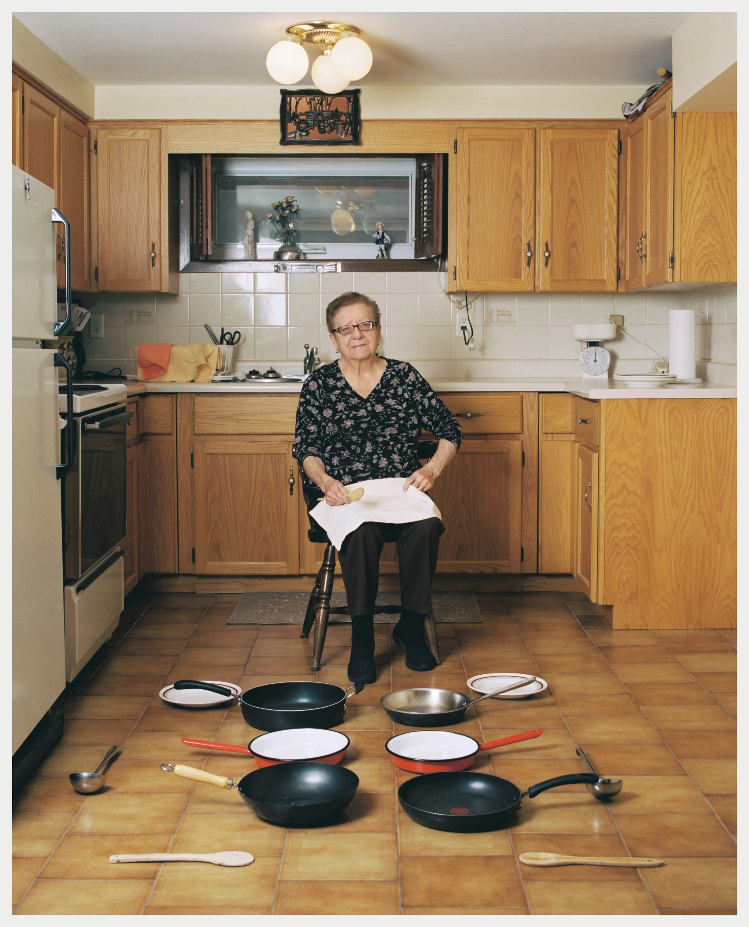 An elderly woman with glasses sitting on a chair in a kitchen, surrounded by multiple frying pans, pots, and cooking utensils on the floor.