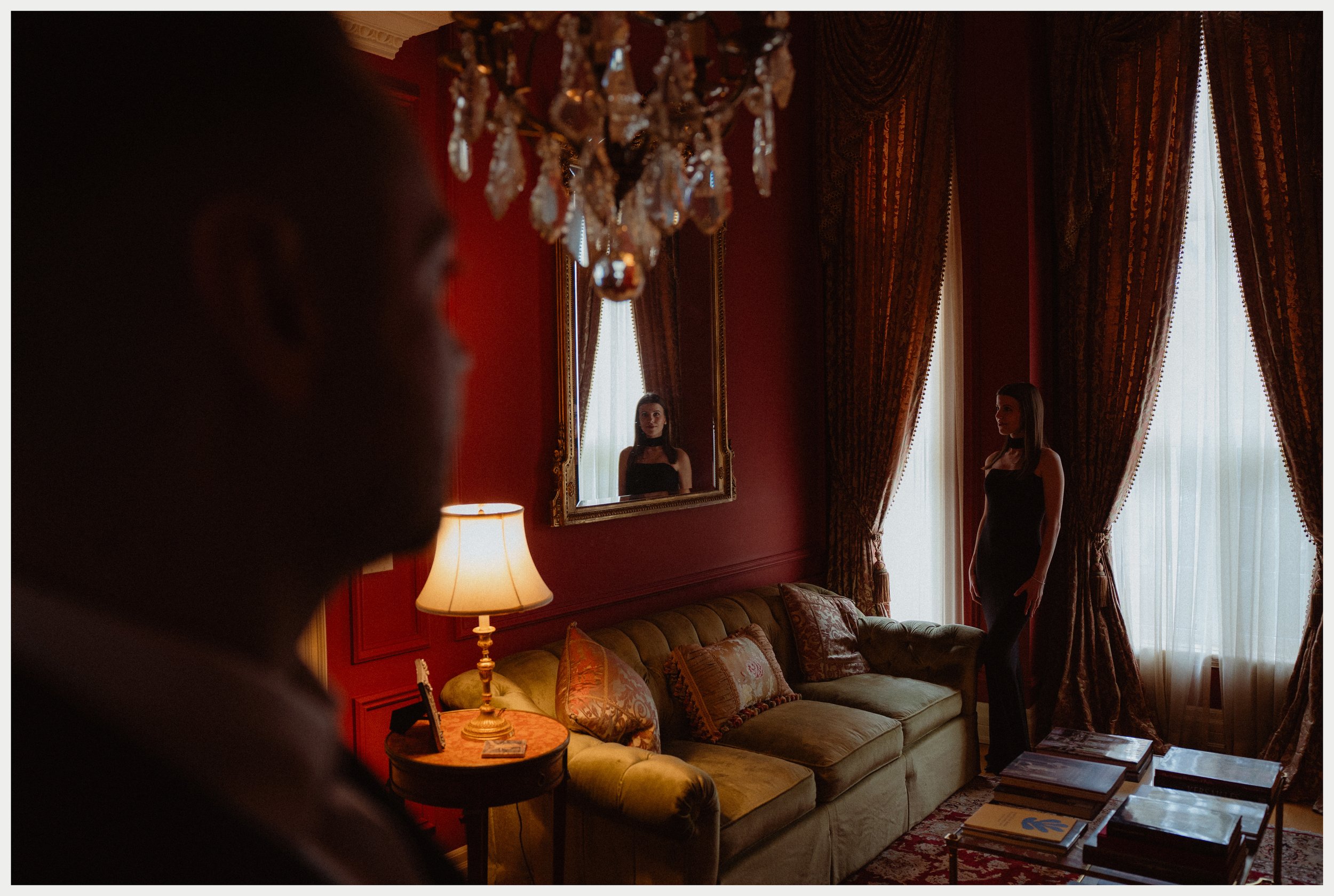 A woman in a black dress standing in a richly decorated room with red walls, gold-framed mirror, chandelier, and heavy curtains, looking at her reflection.