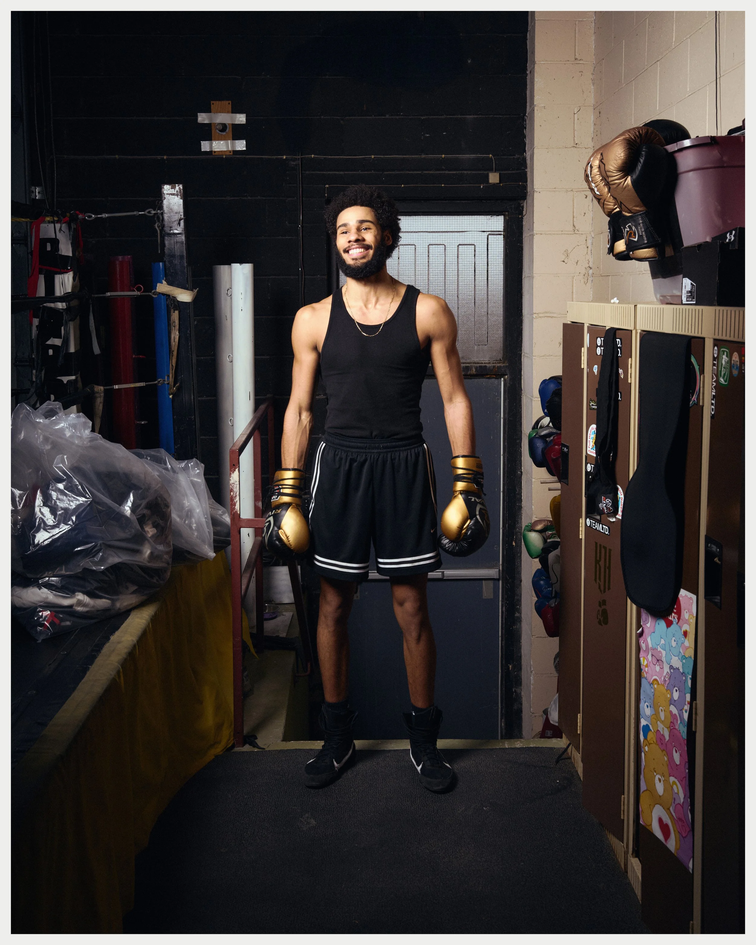 A young man with curly hair and a beard standing in a boxing gym, wearing a black tank top, shorts, and boxing gloves, smiling at the camera. The gym has various boxing equipment and storage shelves around him.