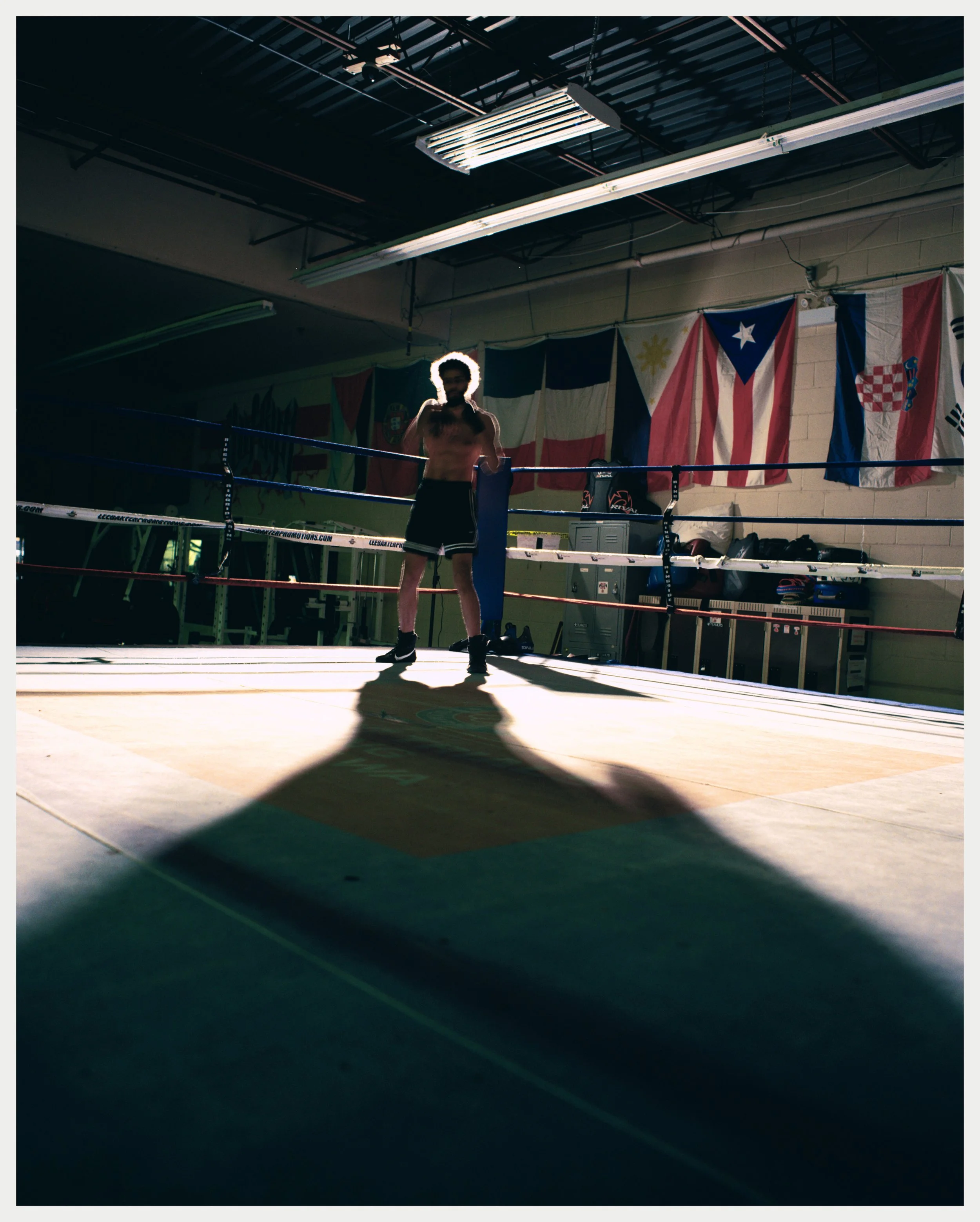 A person standing inside a boxing ring in a gym, illuminated from behind, with flags hanging on the wall behind them.