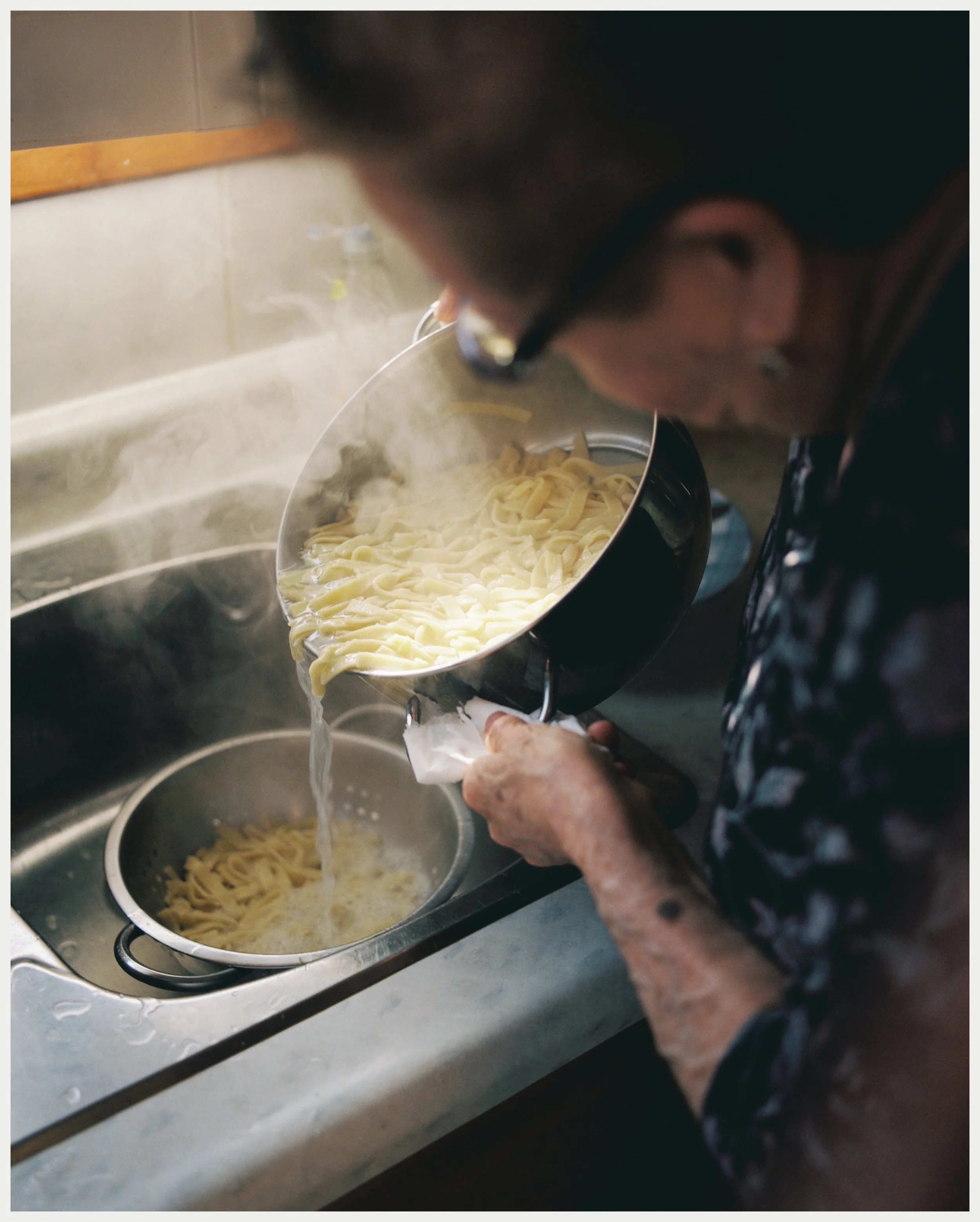 Close-up of an elderly person's hands holding a strainer filled with cooked pasta over a kitchen sink, with steam rising from the pasta.