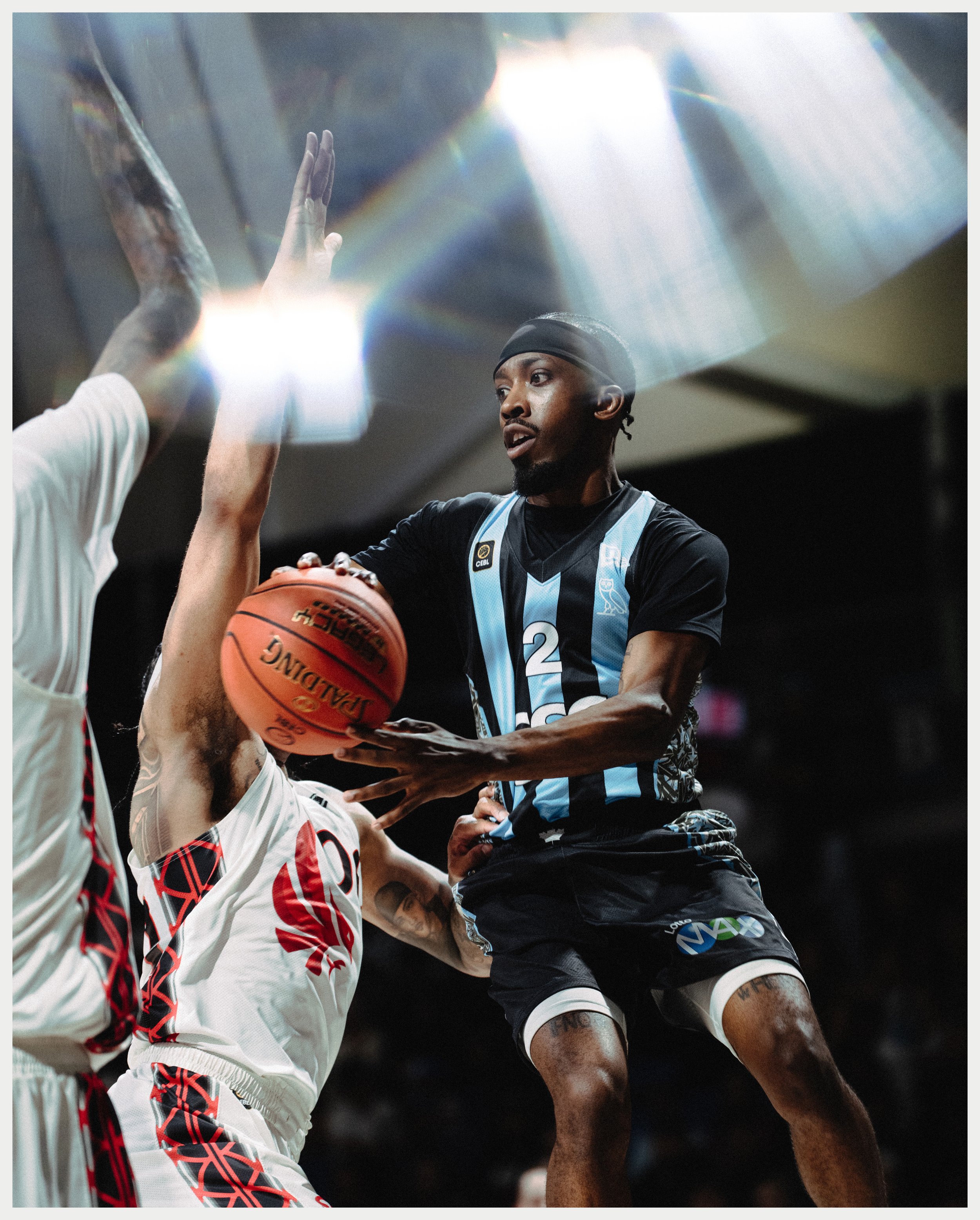 A basketball game featuring two players with high energy and focus, one in a black and blue uniform and the other in a white and red uniform, with a bright light overhead illuminating the scene.