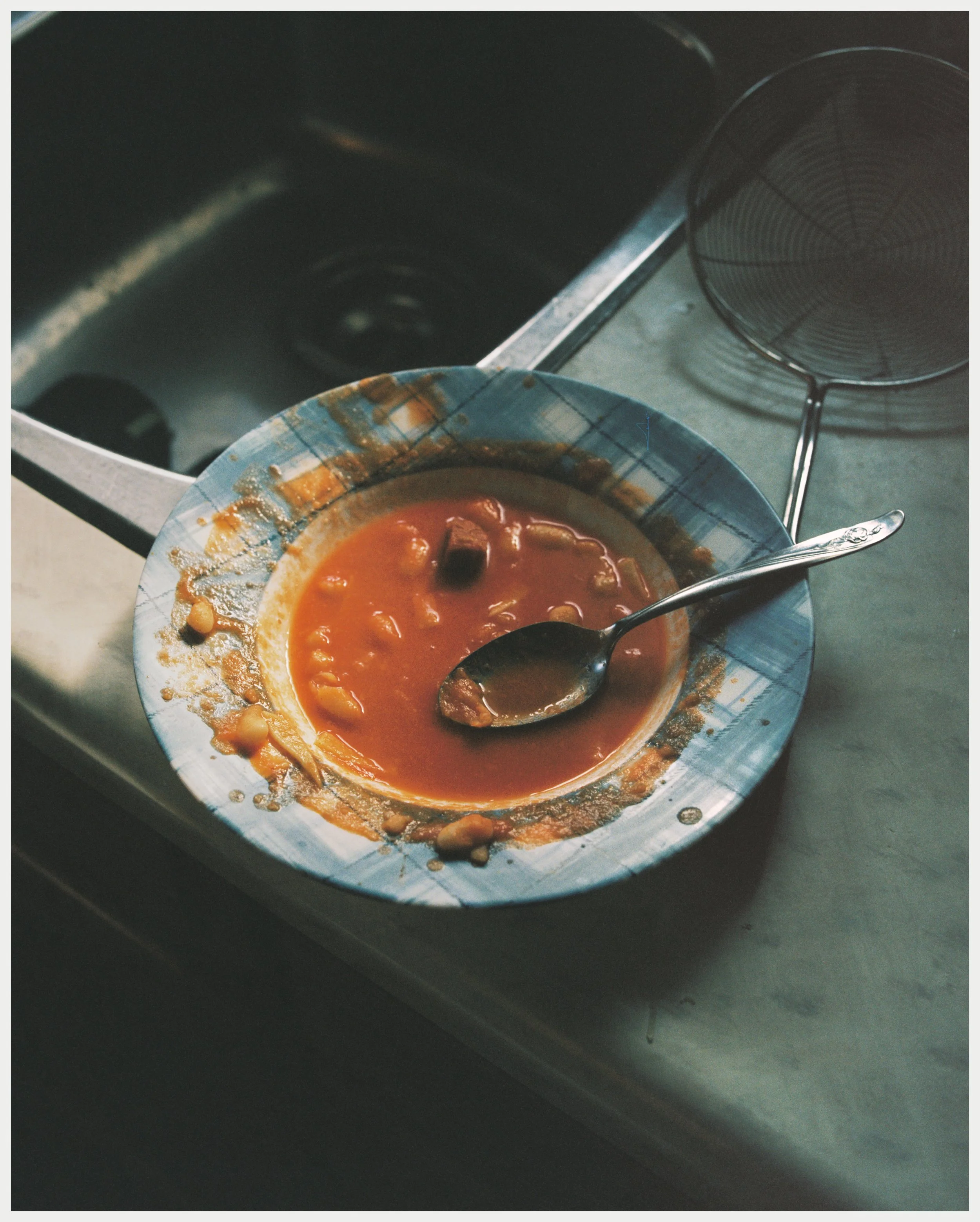 A bowl of tomato-based soup with beans and pieces of meat, on a kitchen counter. A metal spoon rests in the bowl. A metal drying rack is nearby, and a stovetop is in the background.