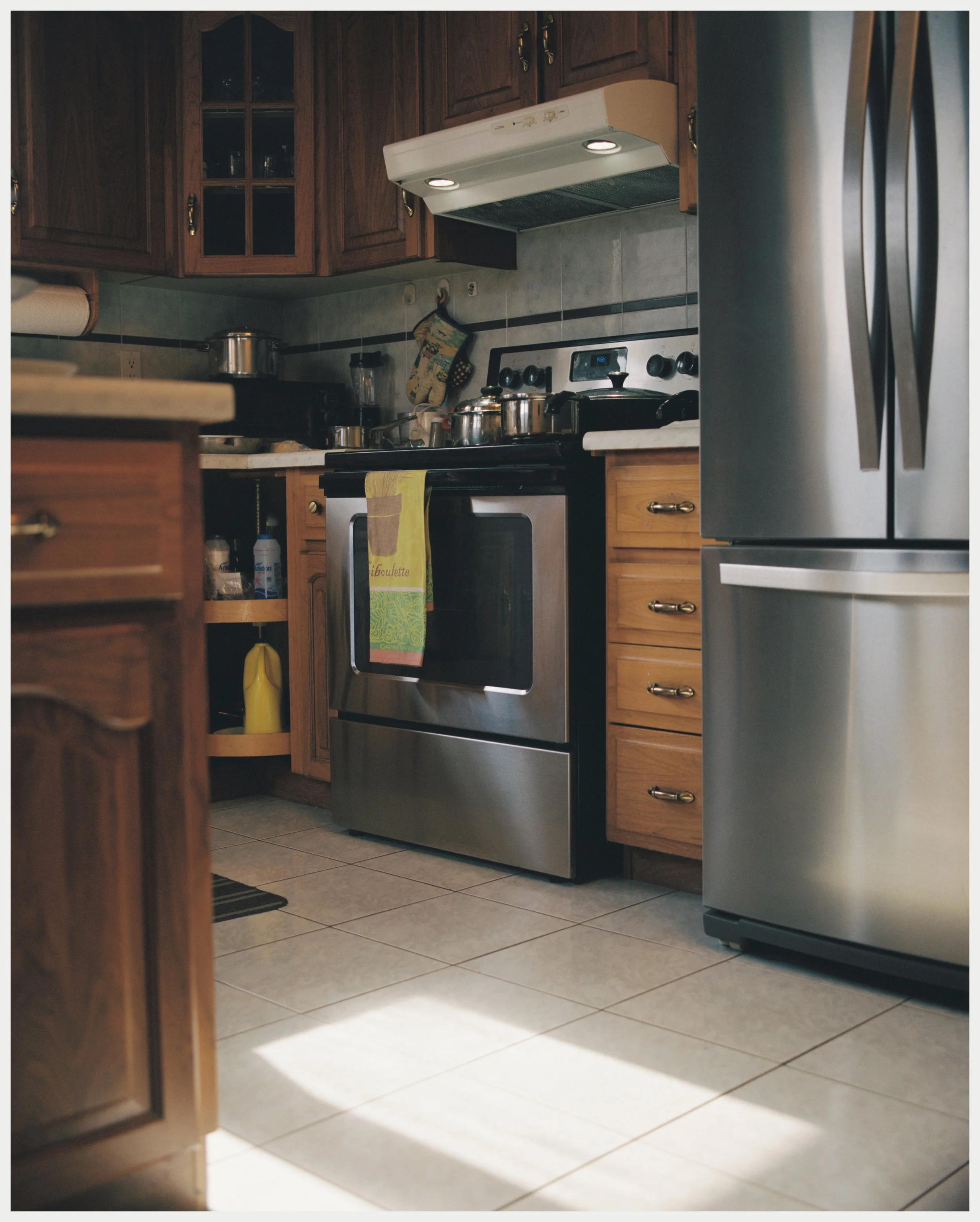 View of a kitchen with wooden cabinetry, stainless steel refrigerator, and a stove with knives and kitchen items on the counter. There are tiled floors and a dish towel hanging on the oven handle.