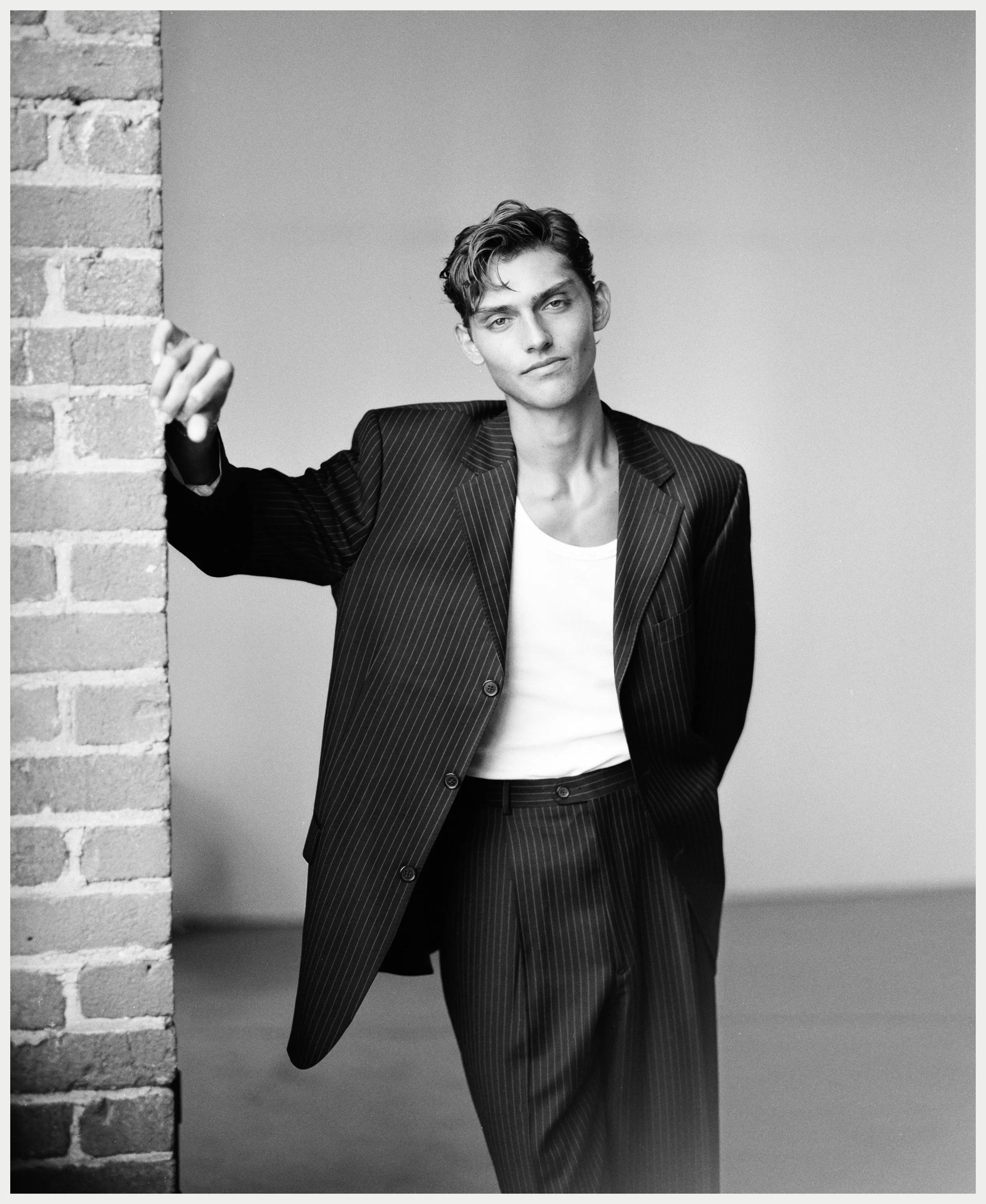 A young man with wavy hair, dressed in a pinstripe blazer and white shirt, leaning against a brick wall with one hand, looking at the camera with a confident expression, in a black-and-white photo.