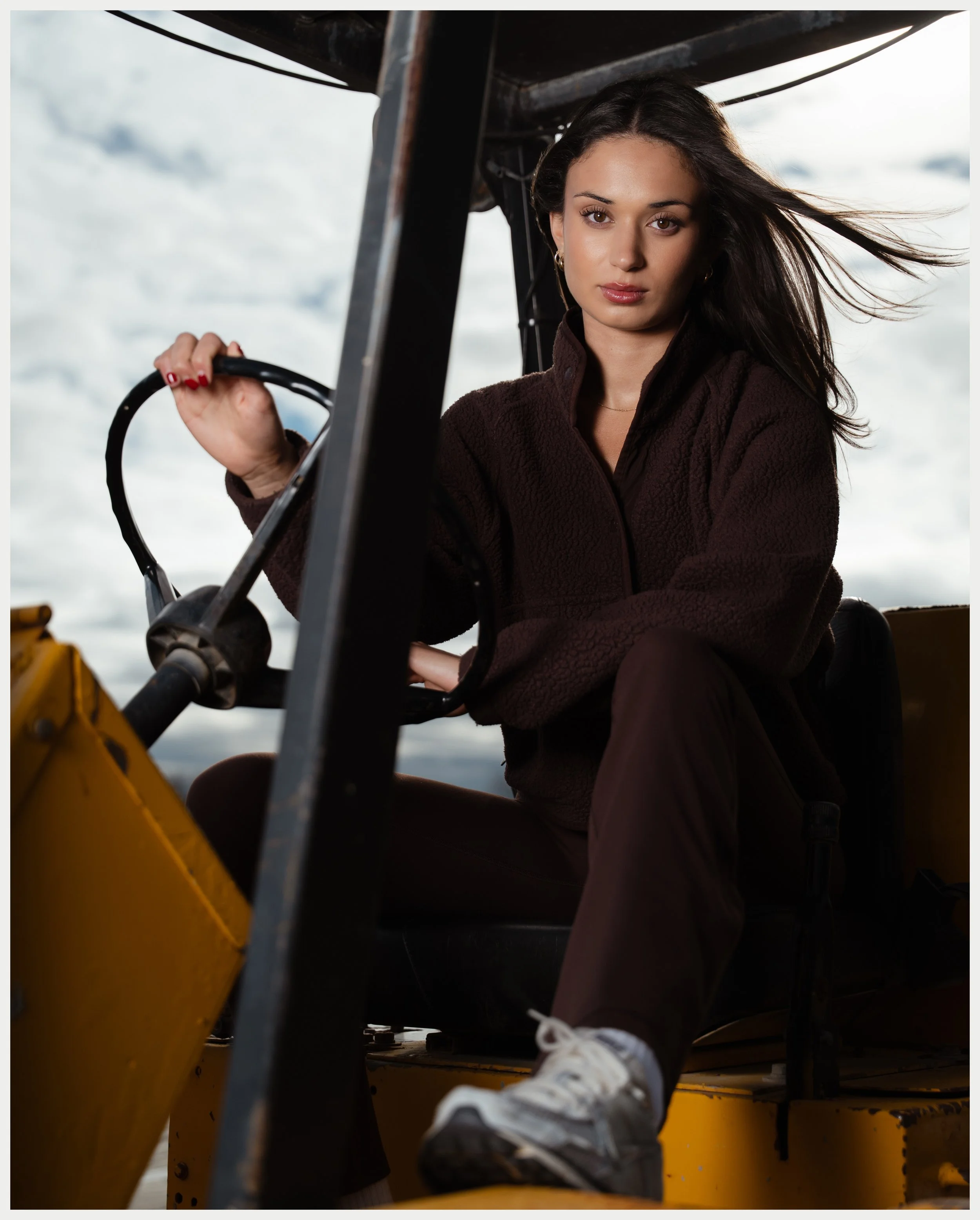 A woman with dark hair, wearing a brown jacket and white sneakers, sitting in a yellow boat, holding a control handle, with a cloudy sky in the background.