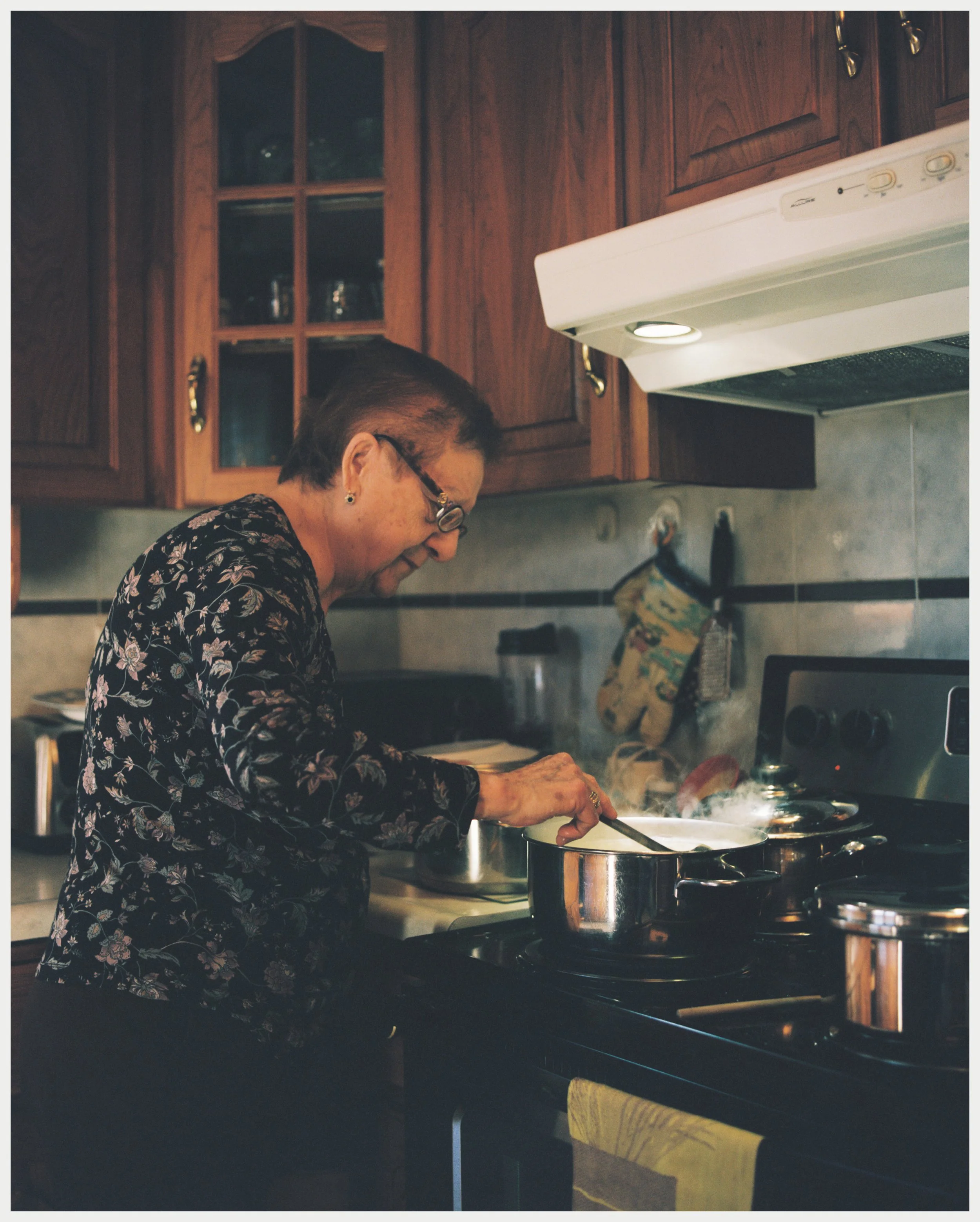 An elderly woman with glasses and earrings, wearing a floral top, cooking on a stove in a kitchen with wooden cabinets and a yellow towel on a chair in the foreground.