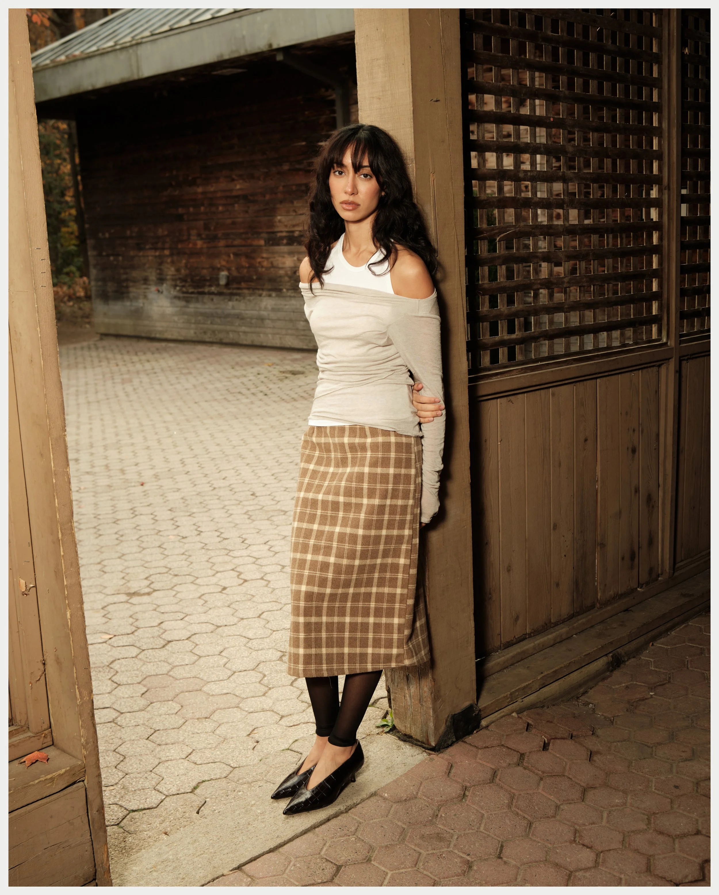 A woman with dark wavy hair leaning against a wooden post between a wooden fence and an open gate, wearing a beige slash-shoulder top, plaid skirt, black tights, and black heels, standing on a stone pathway outside an area with a brick wall.