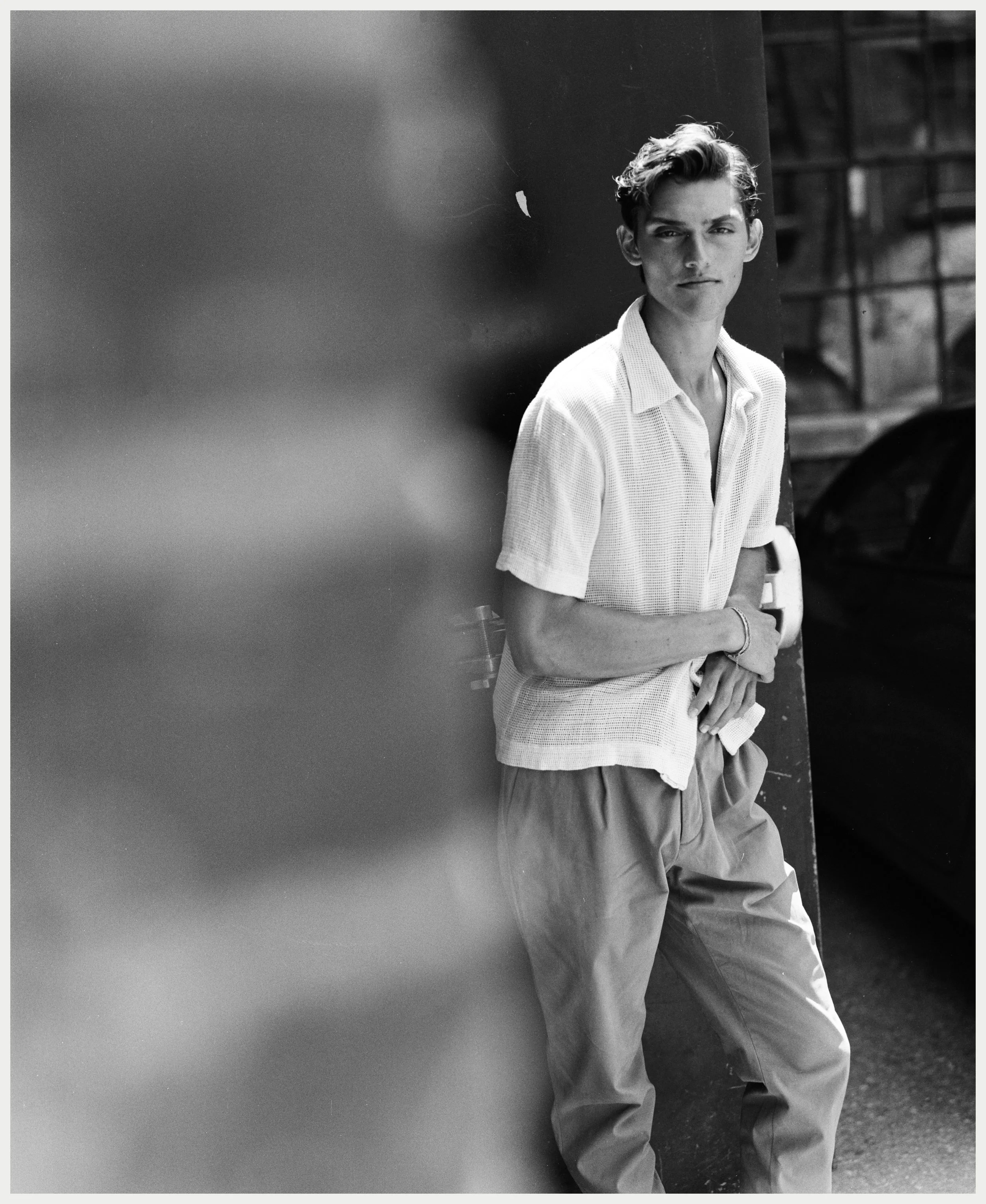 A black and white photograph of a young man with styled hair and a serious expression, standing outdoors against a background with a dark wall or structure. He is wearing a light-colored short-sleeve shirt and baggy pants.