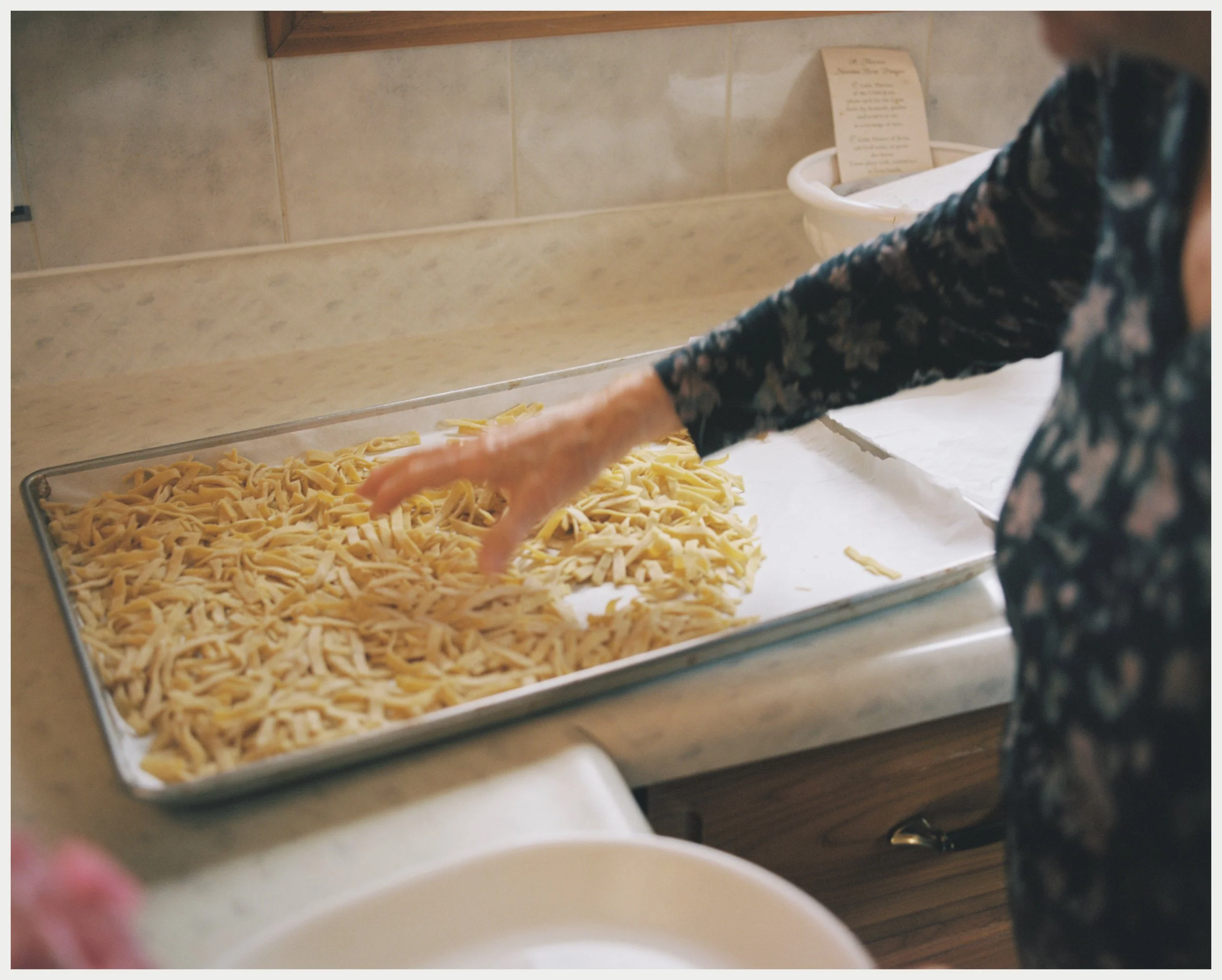 A person with a long-sleeved floral shirt is reaching towards a tray of uncooked fettuccine pasta on a kitchen countertop. In the background, there are beige tiled walls and a white basket.