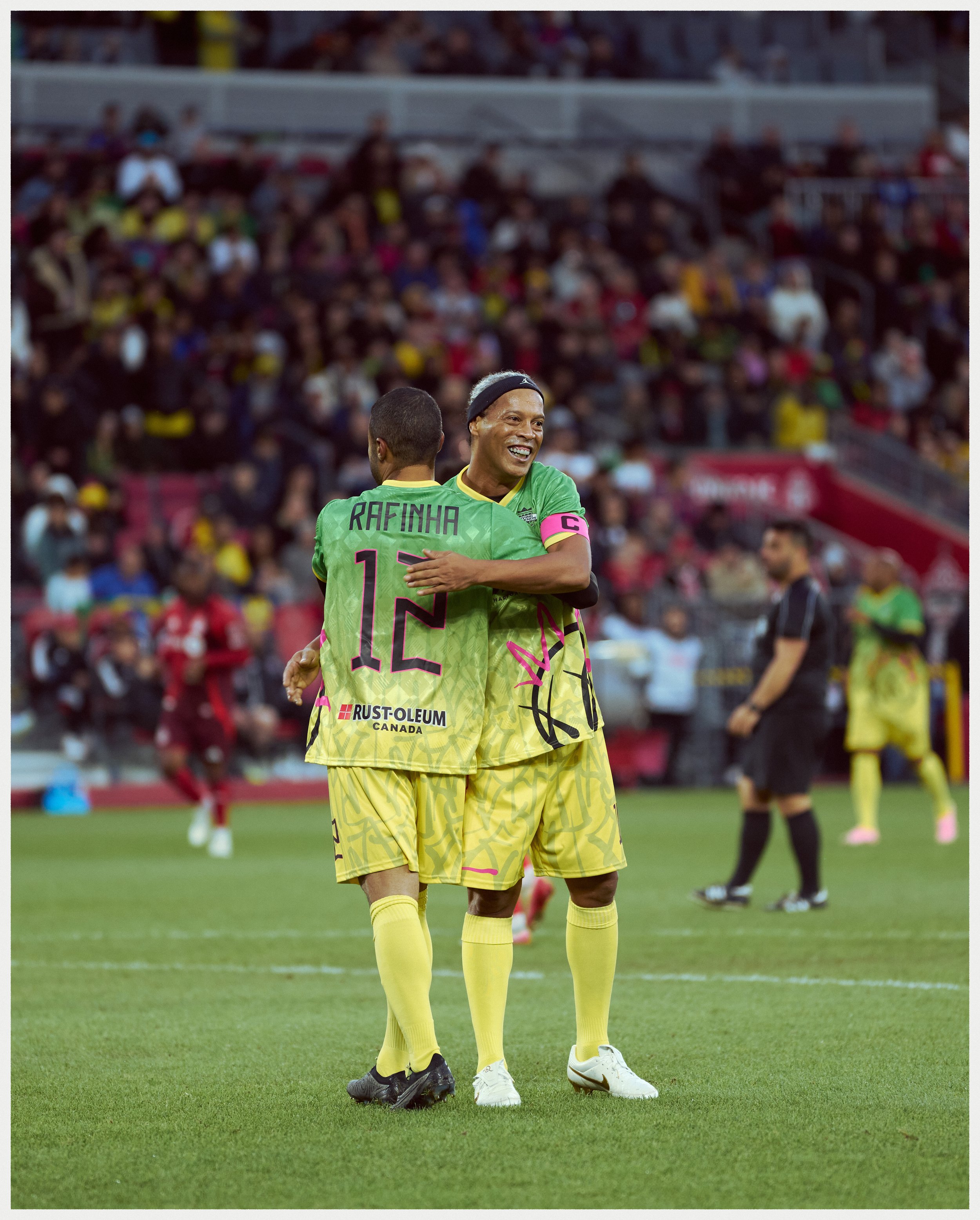 Two soccer players in yellow and green uniforms hugging on a field during a match, with a large crowd in the stands in the background.