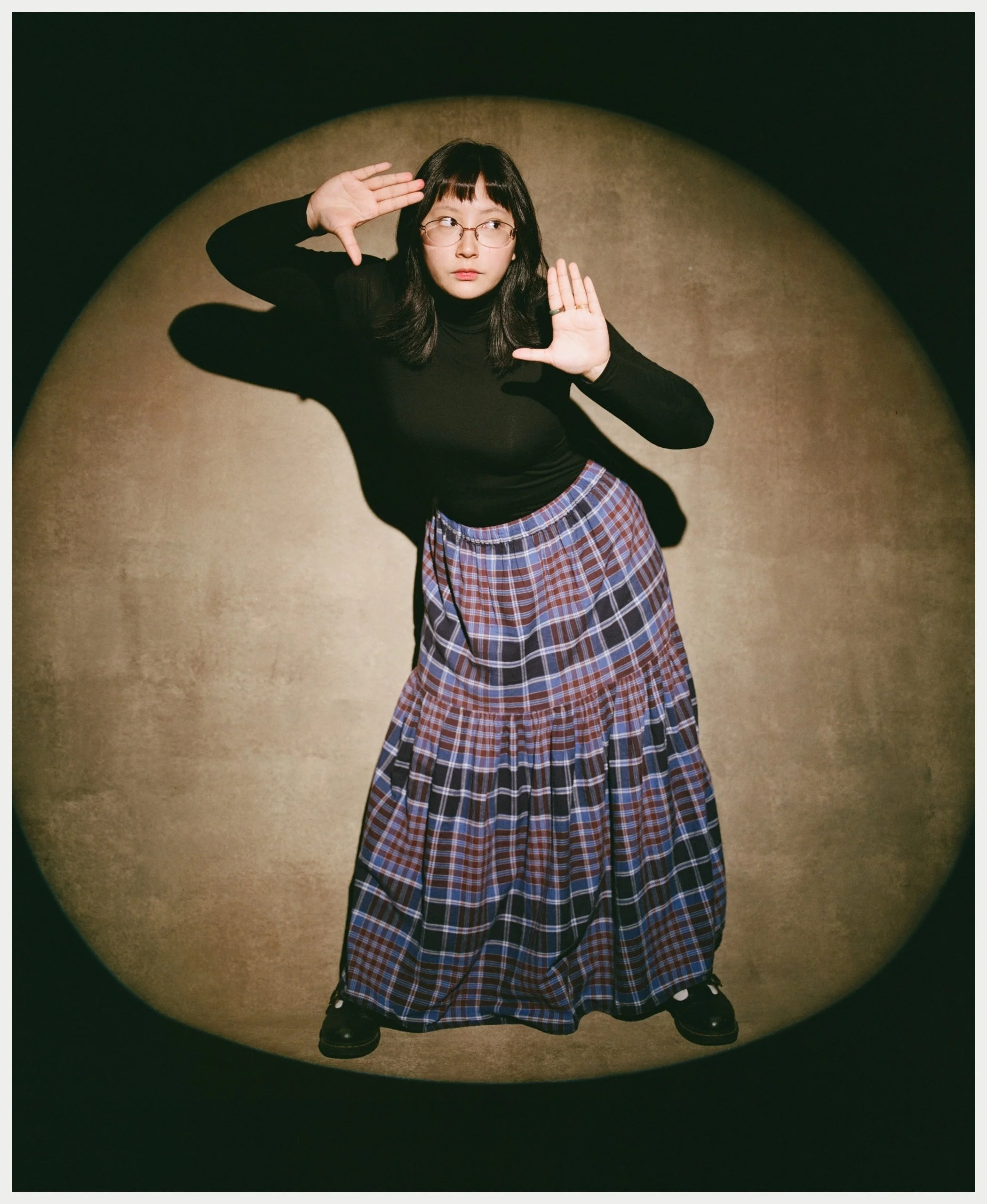 A woman with black hair, glasses, and a black long-sleeve top is posing with her hands raised near her face, standing on a beige-colored carpet in front of a dark background. Her long plaid skirt reaches her ankles, and she wears black shoes.