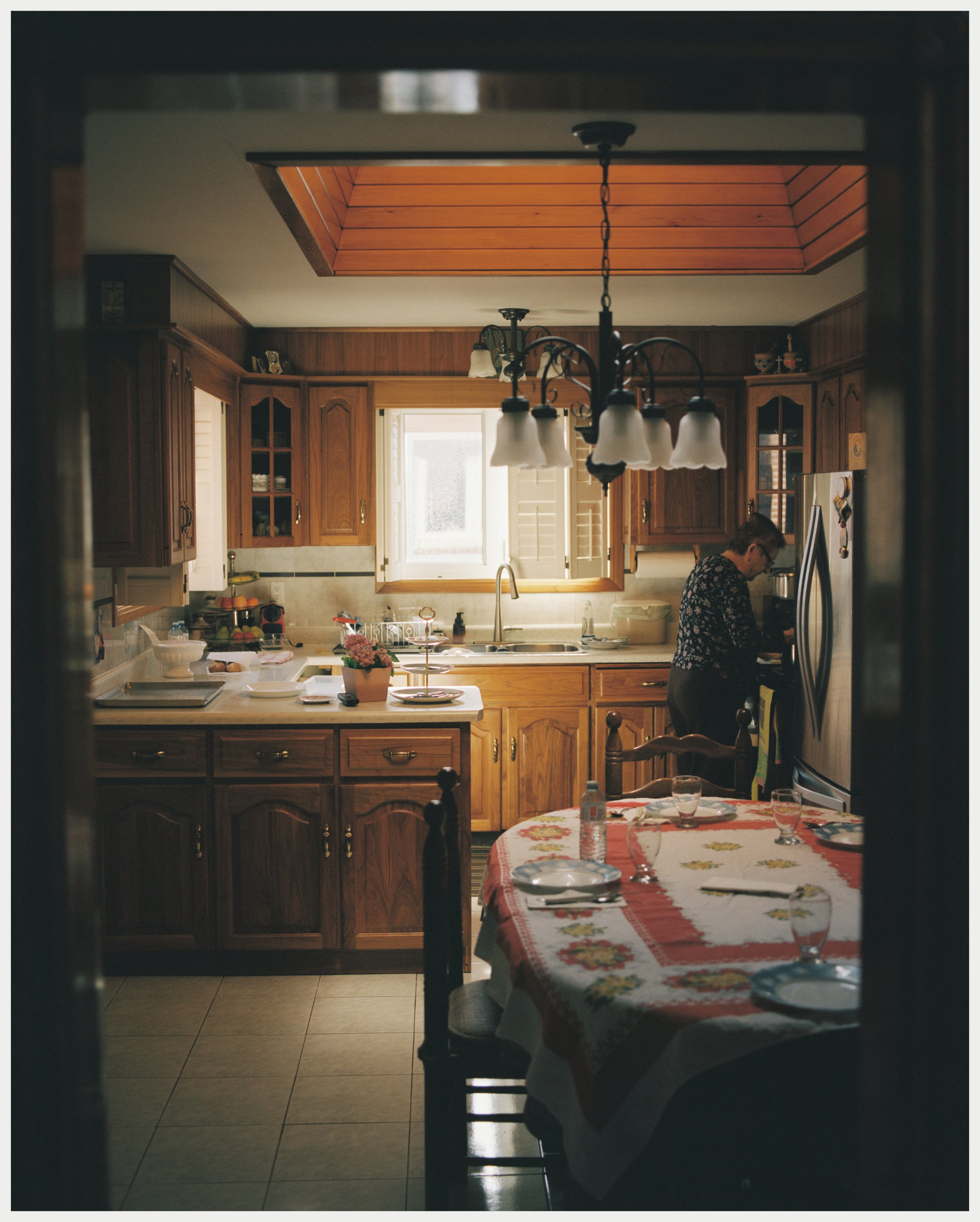 View of a kitchen with wooden cabinets, a window, and a woman preparing food in front of a refrigerator, through an open doorway with a dining table covered with a red floral tablecloth in the foreground.