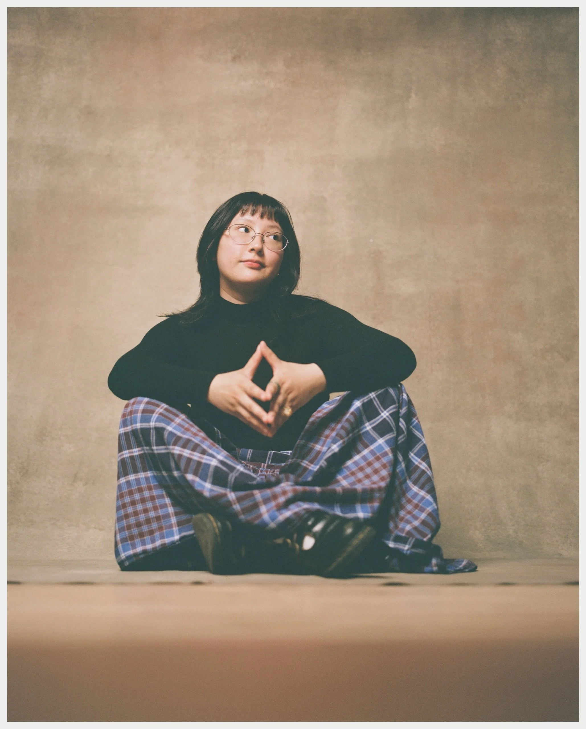 A woman with shoulder-length dark hair, glasses, and wearing a black long-sleeve top and plaid pants, sitting on the floor in front of a beige background, with her hands together forming a triangle.