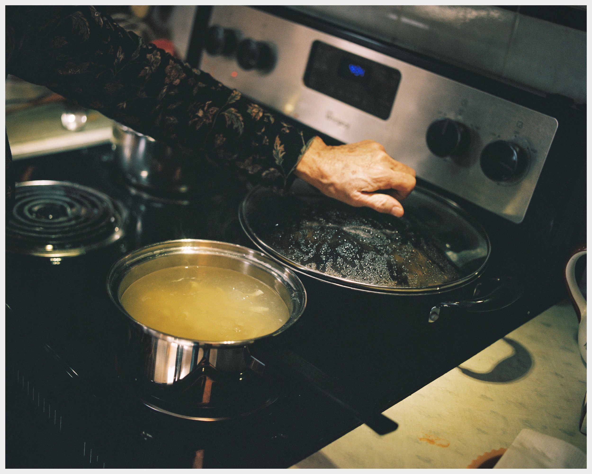 A person's hand reaching into a pot on a stove, with a bowl of what appears to be melted butter or oil nearby. The stove has black burners and a control panel with black knobs and a digital display. The scene suggests a cooking process in a kitchen.