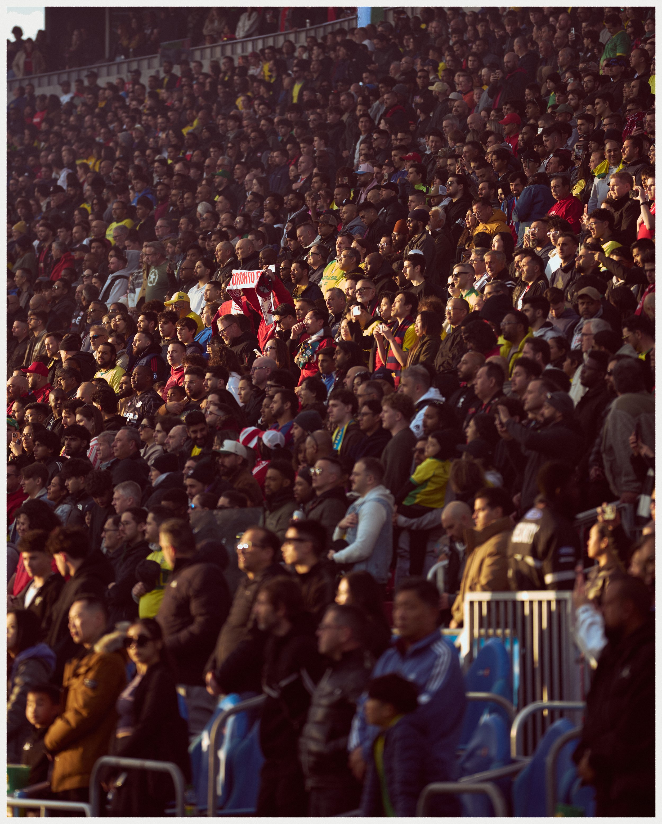 Crowd of people sitting and standing in stadium seats, attending an outdoor sporting event, many wearing colorful clothing and some holding signs.