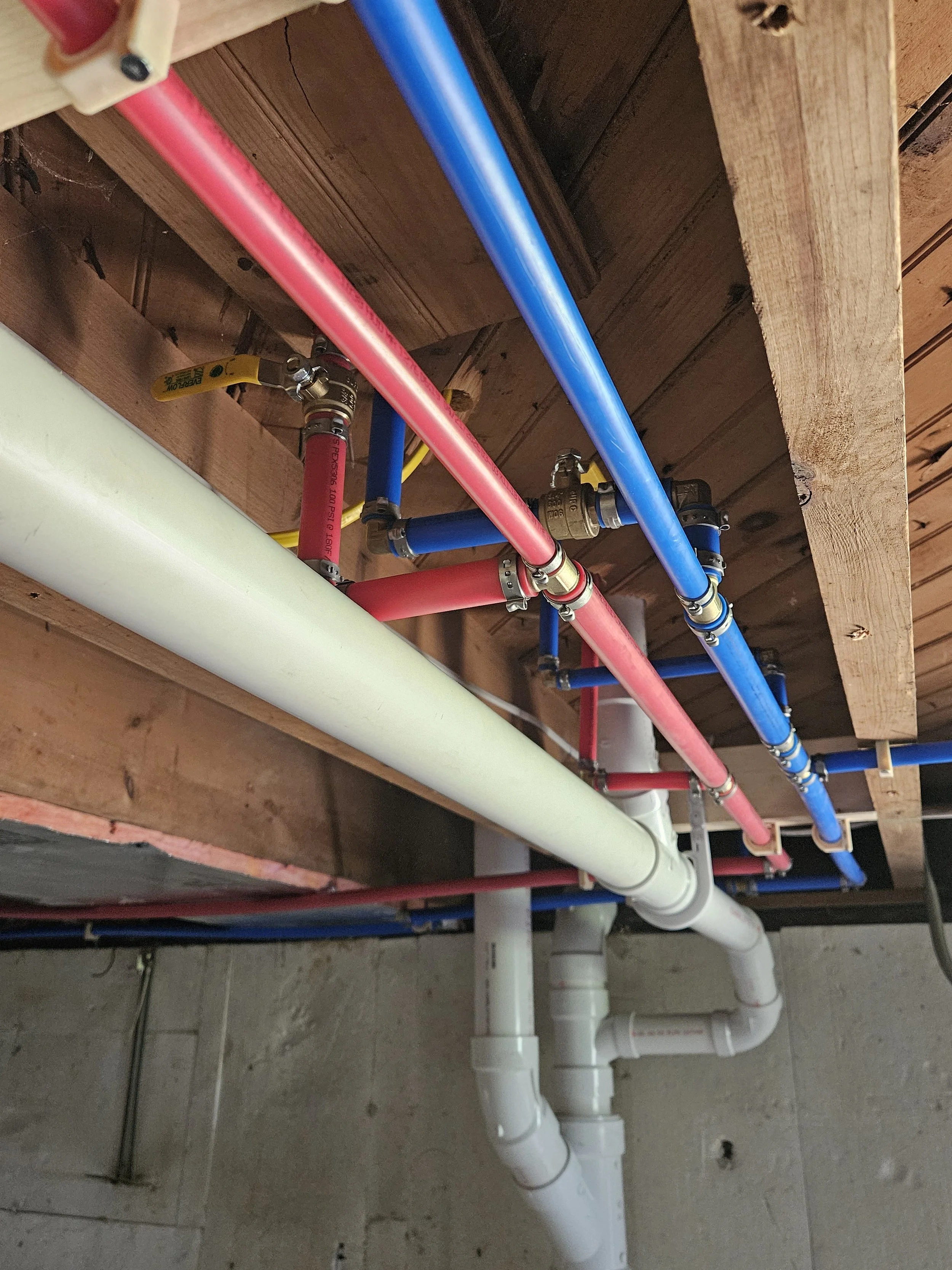 Pipes and plumbing installed underneath a wooden floor or ceiling. Colored pipes include red, blue, yellow, and white, connected with various fittings.