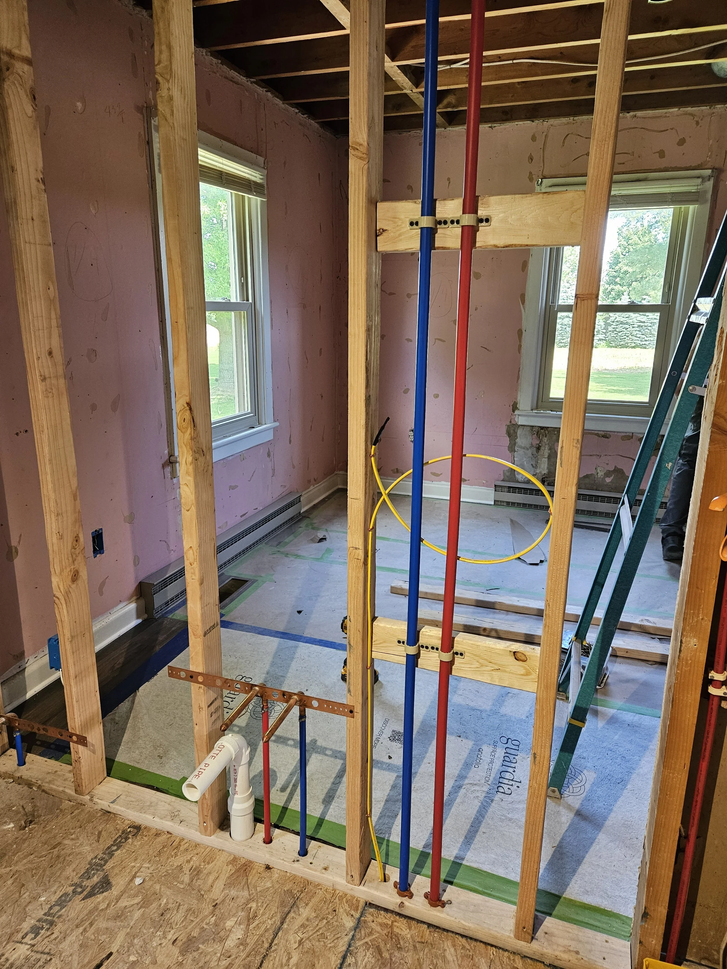 Interior of a house under construction, showing exposed wooden framing, plumbing pipes, and two windows with a view of trees outside.