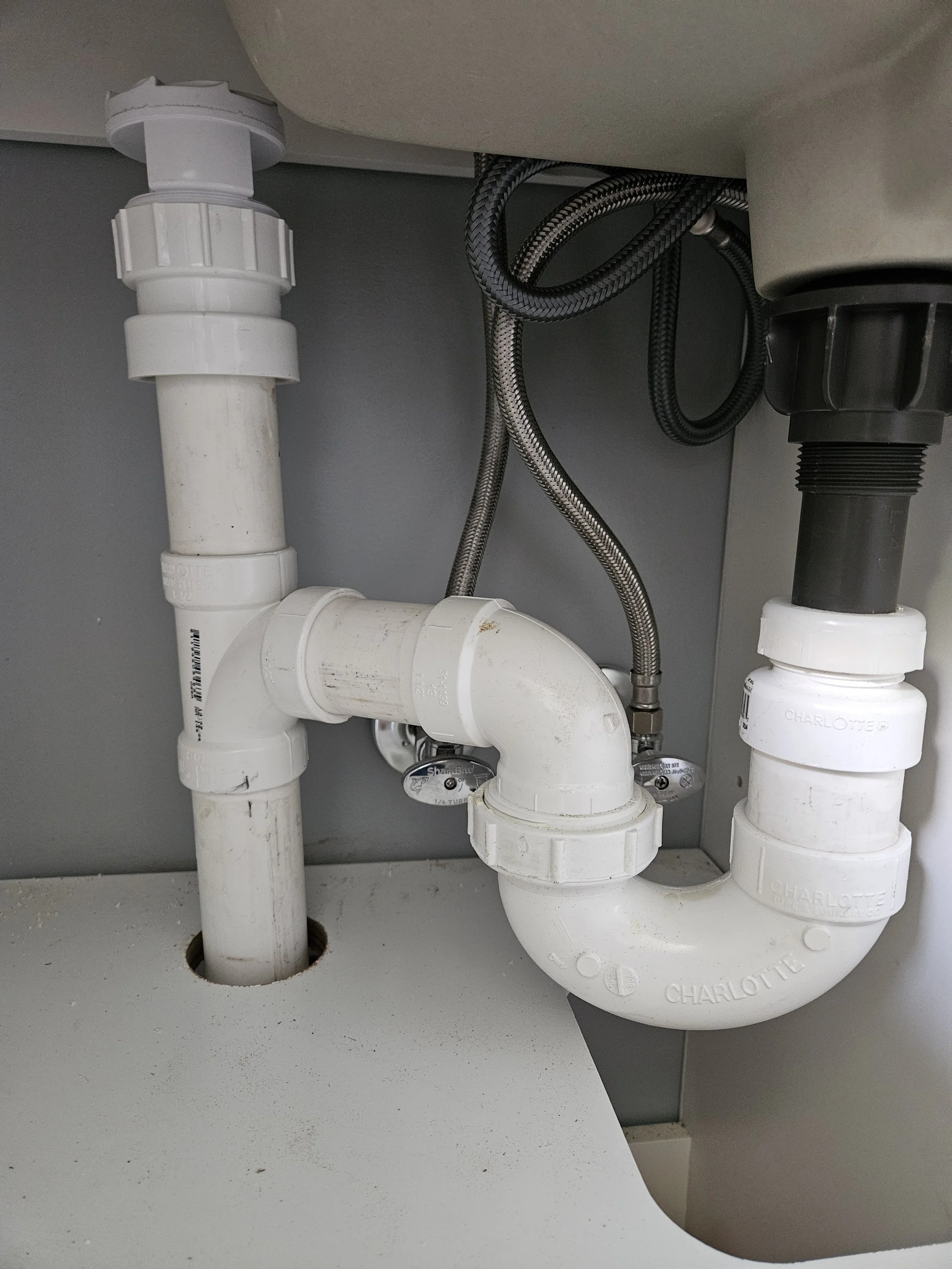 Underneath the kitchen sink, there are white PVC pipes connected to plumbing fixtures. Some flexible metal braided tubing is also visible, connecting to the black drain pipe on the right side.