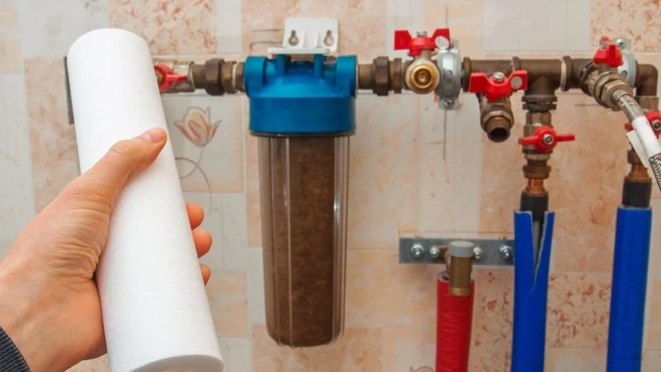 A person holding a water filter cartridge near plumbing pipes and valves on a tiled wall.