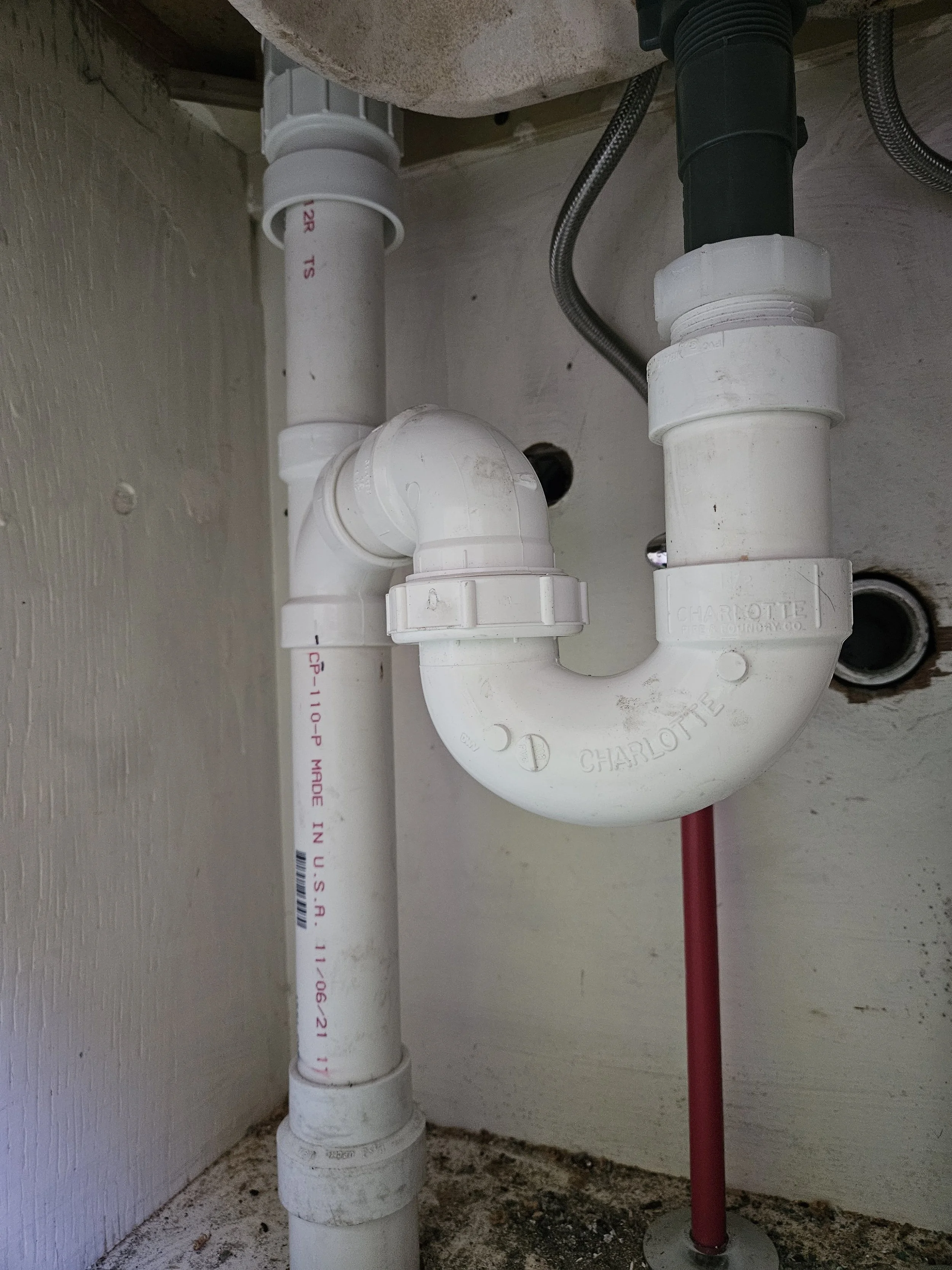 Underneath a sink showing various plumbing pipes, including PVC pipes, a black pipe, a silver braided pipe, and a red pipe on a concrete floor with some dirt.