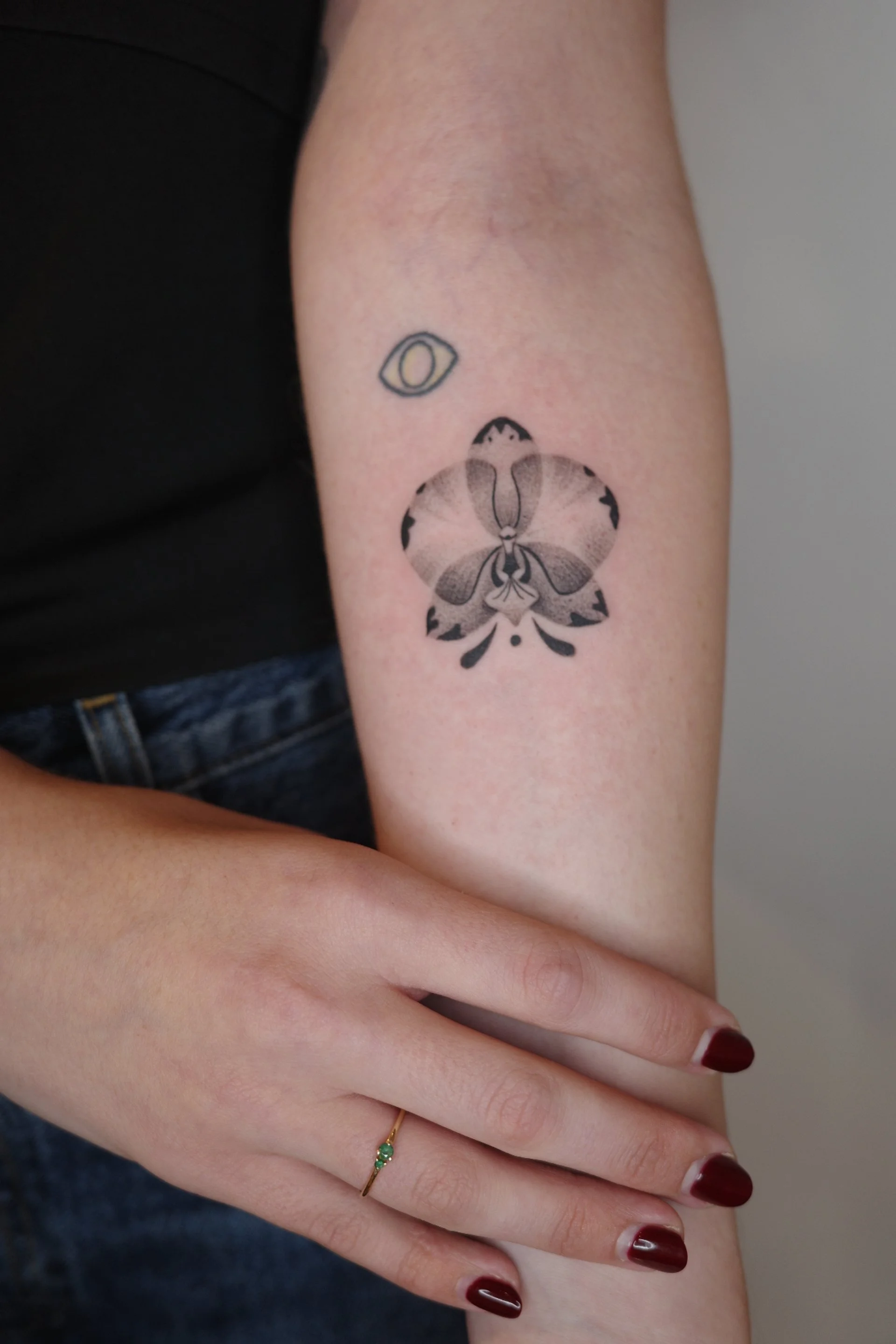 A person's arm with tattoos and jewelry, including a black and gray moth tattoo and a small, colored eye tattoo, resting on their lap with nails painted dark red.