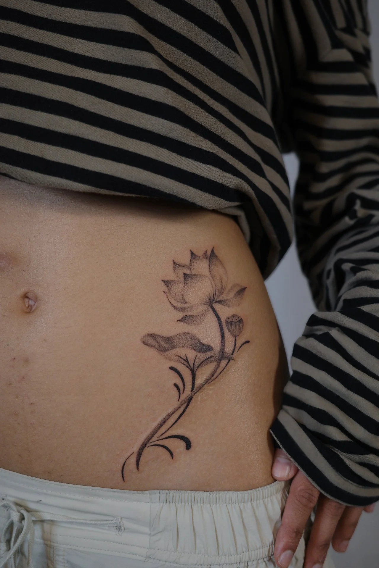 Close-up of a person's side abdomen with a black and gray flower tattoo, wearing a black and white striped shirt and beige pants.