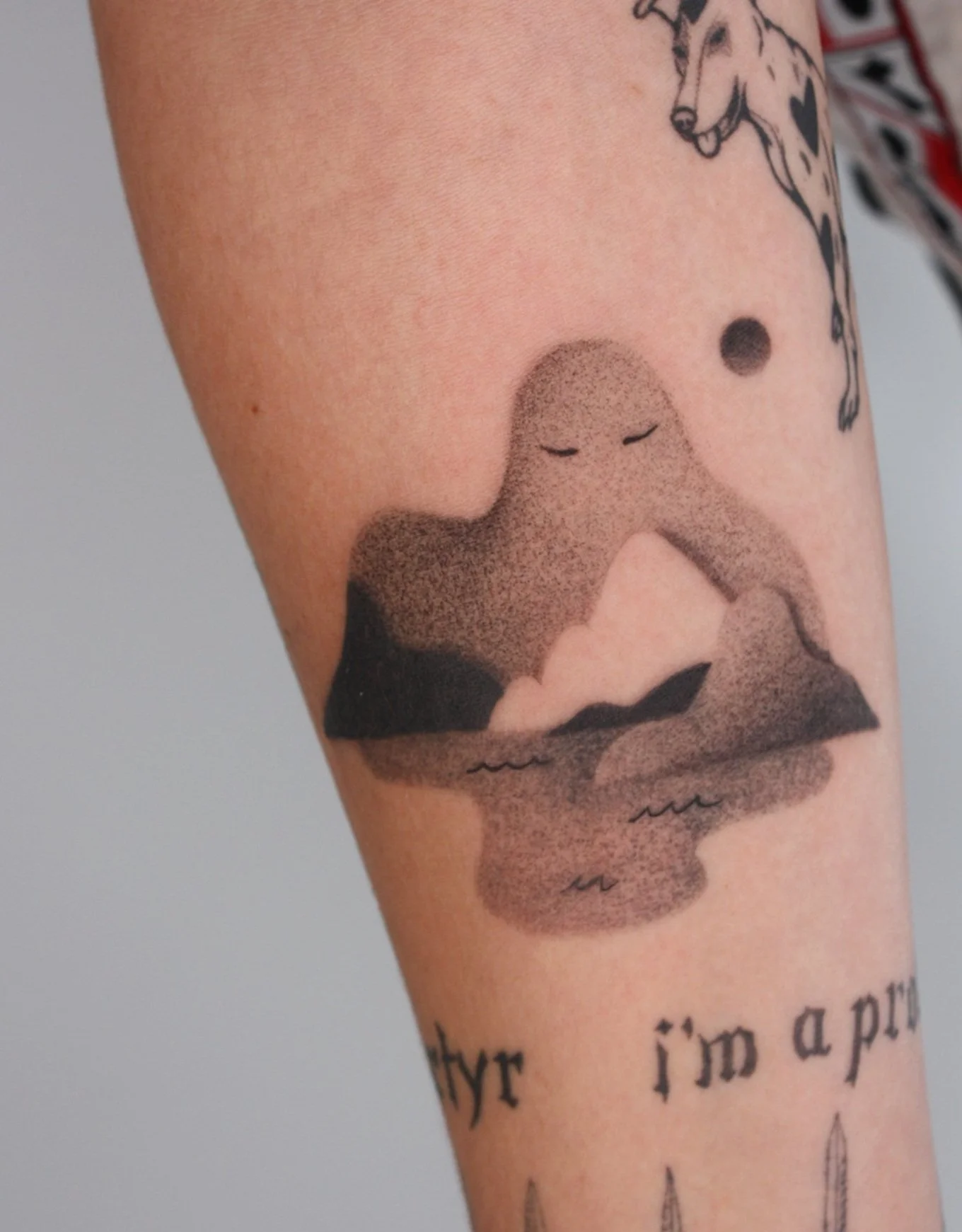 Tattoo of a smiling mountain with a face, clouds, and water below on a person's forearm.
