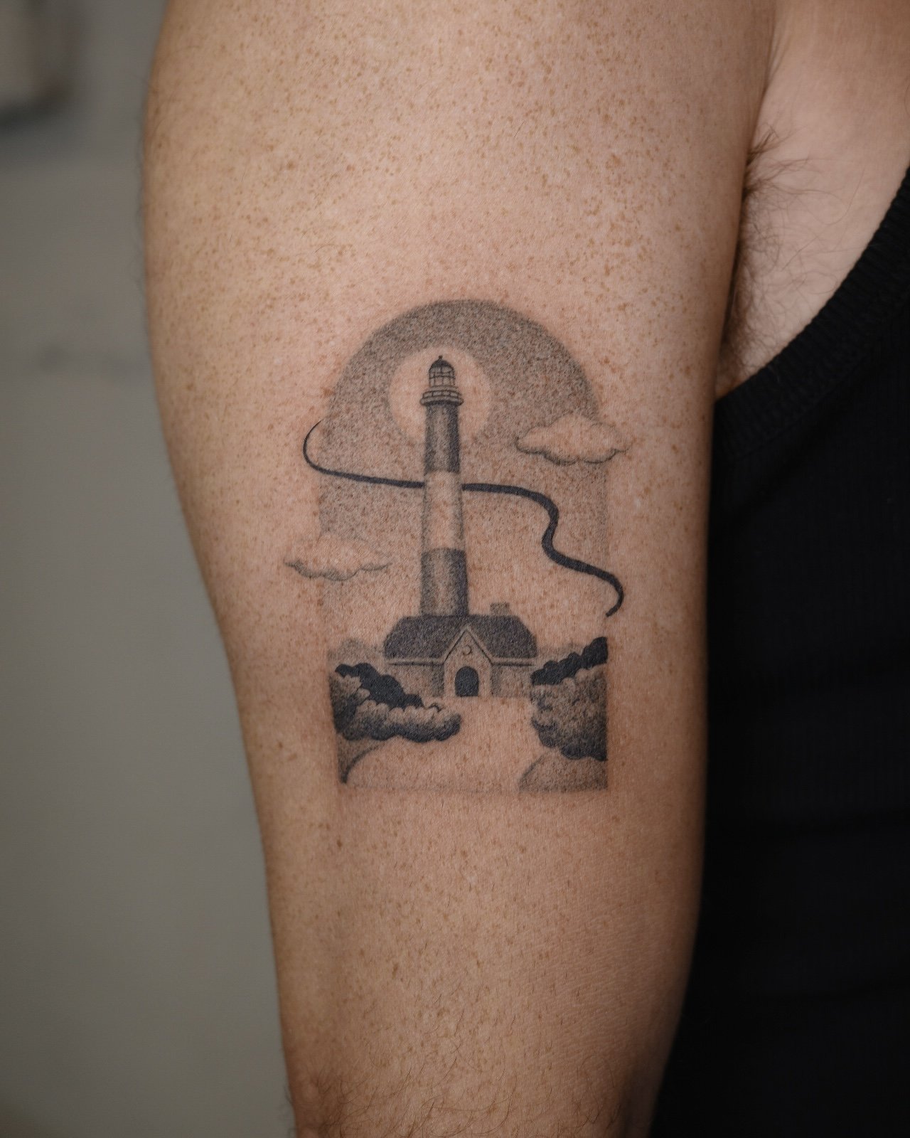 Tattoo of a lighthouse on person's upper arm with clouds and a curved path.