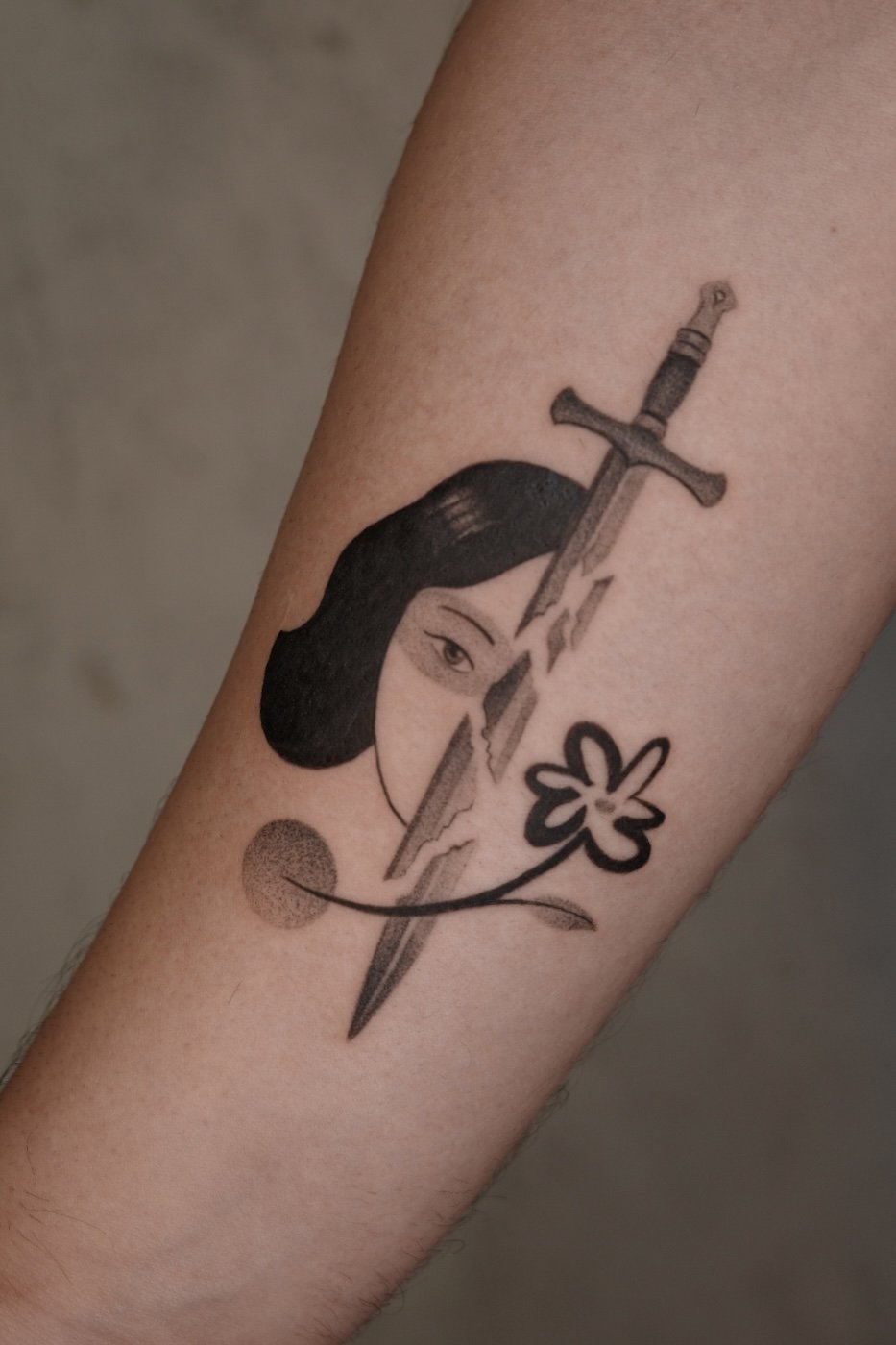 Tattoo of a woman's face with dark hair, a sword through the forehead, a flower, and a small circle.