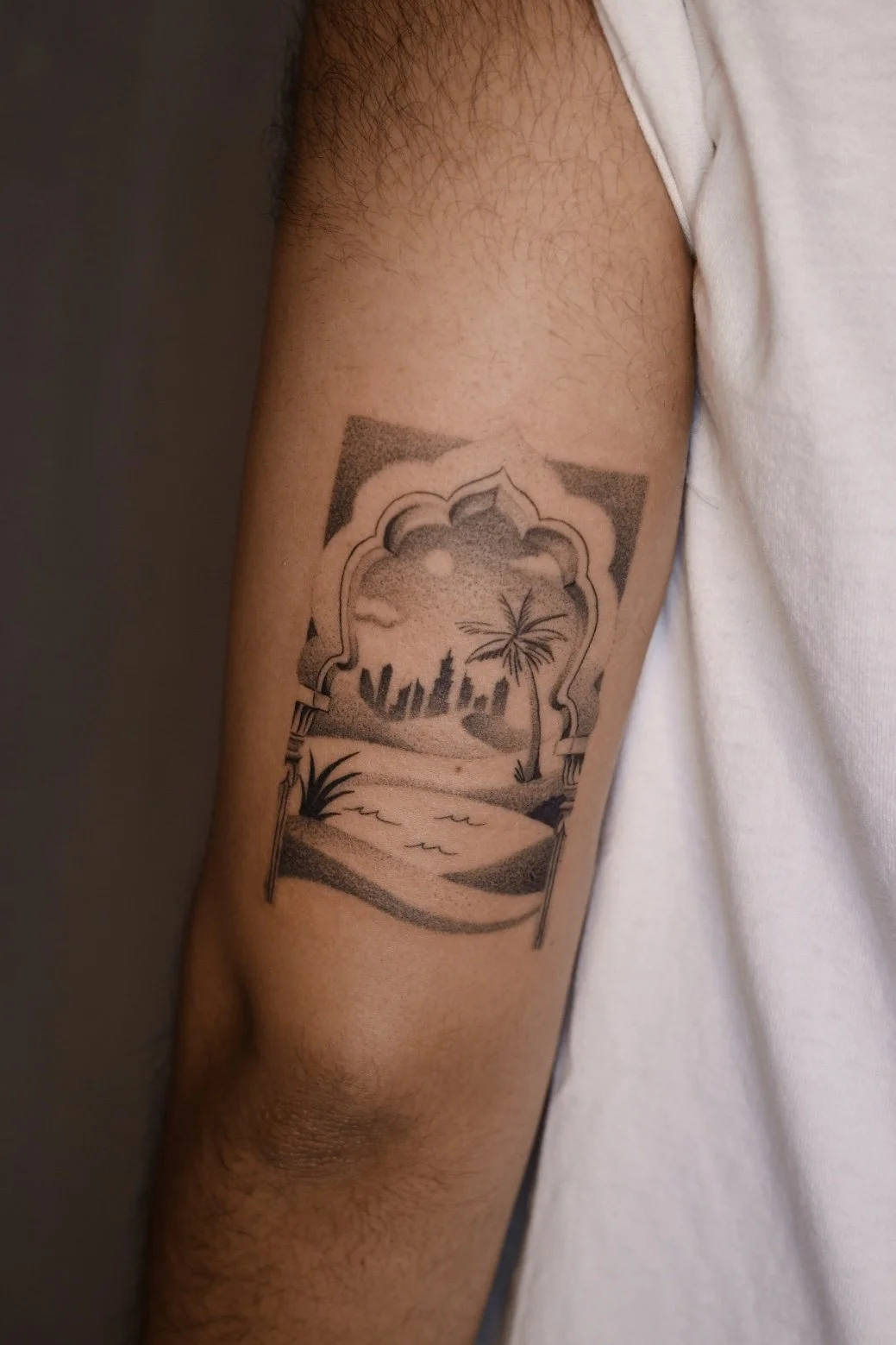 A tattoo on a person's upper arm shows a city skyline, a large palm tree, and a beach scene framed by an ornate archway.