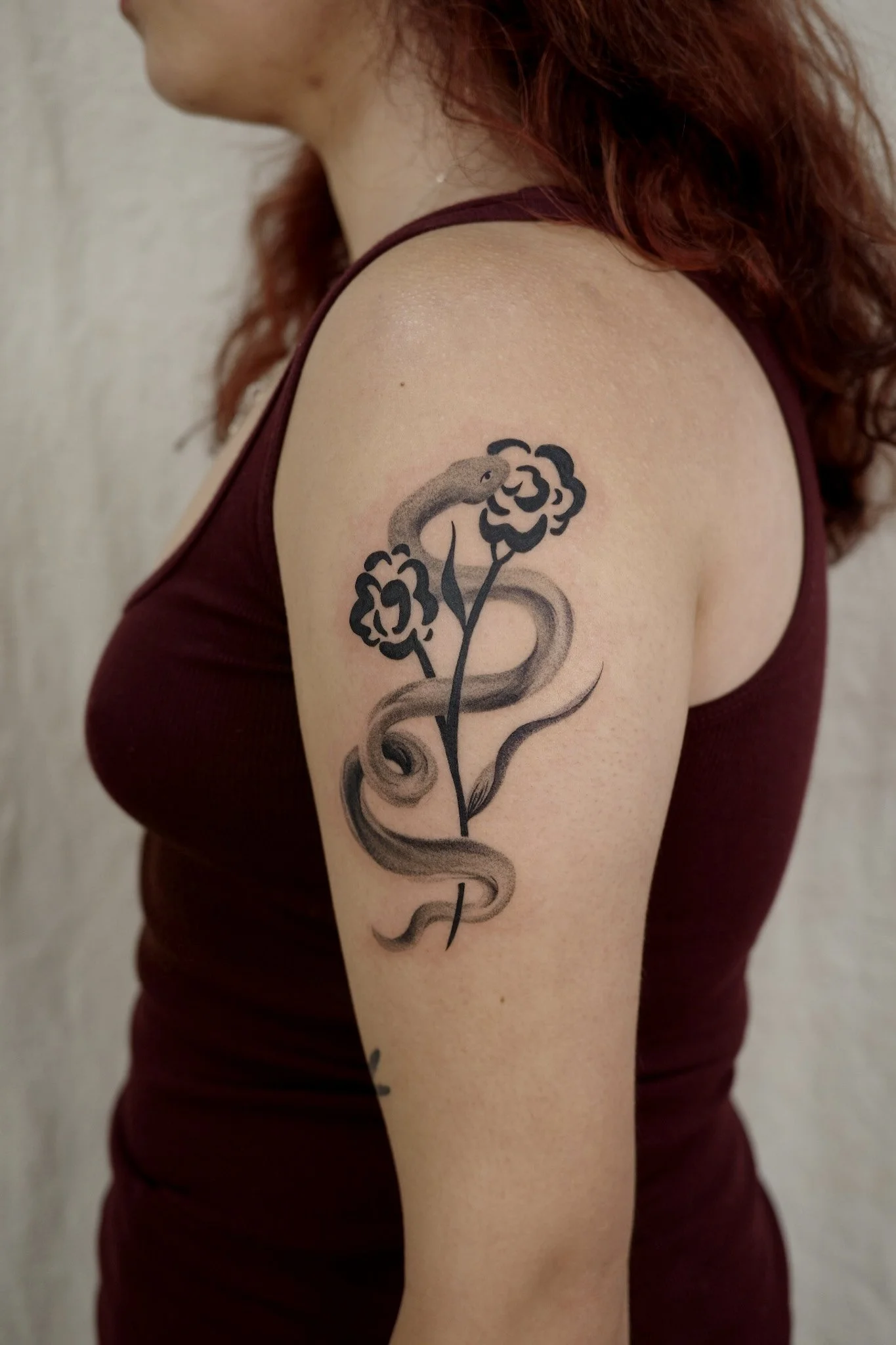 A woman with red hair wearing a sleeveless maroon top, showing a black and gray tattoo of a snake and a flower on her upper arm.