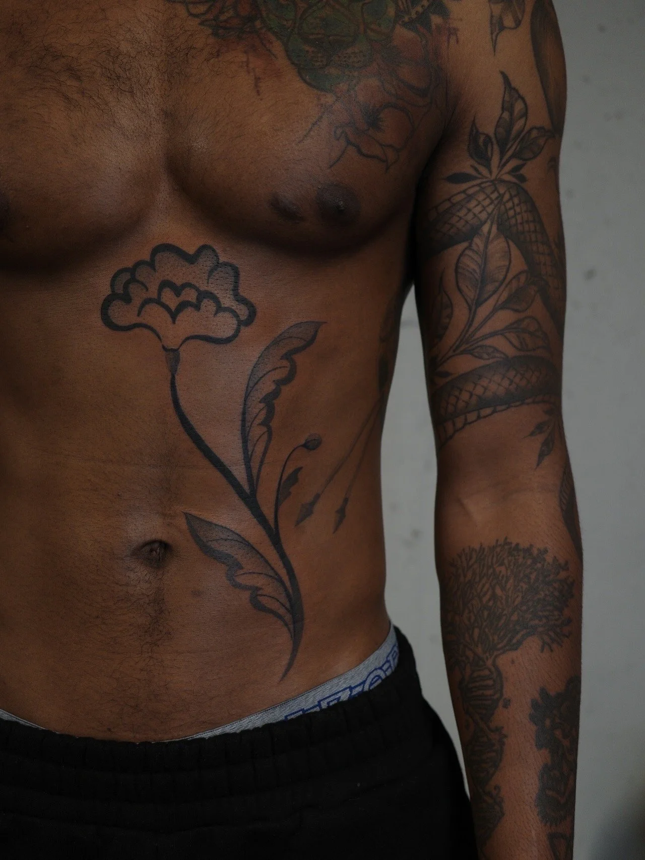 Close-up of a shirtless man with multiple tattoos on his chest and right arm, wearing black shorts.