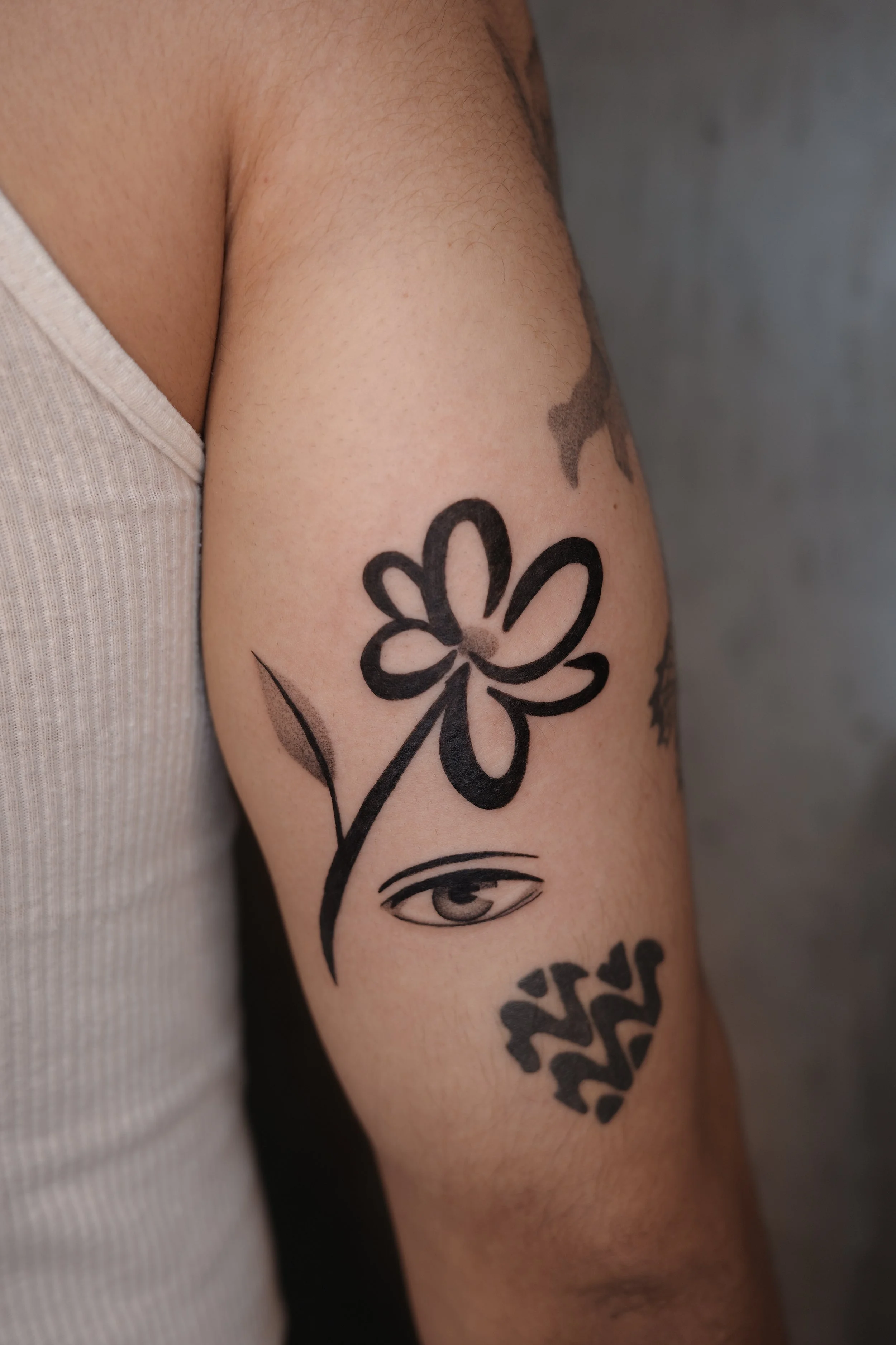 Close-up of a person's upper arm with tattoos, including a flower design, an eye, and text, with a gray background.