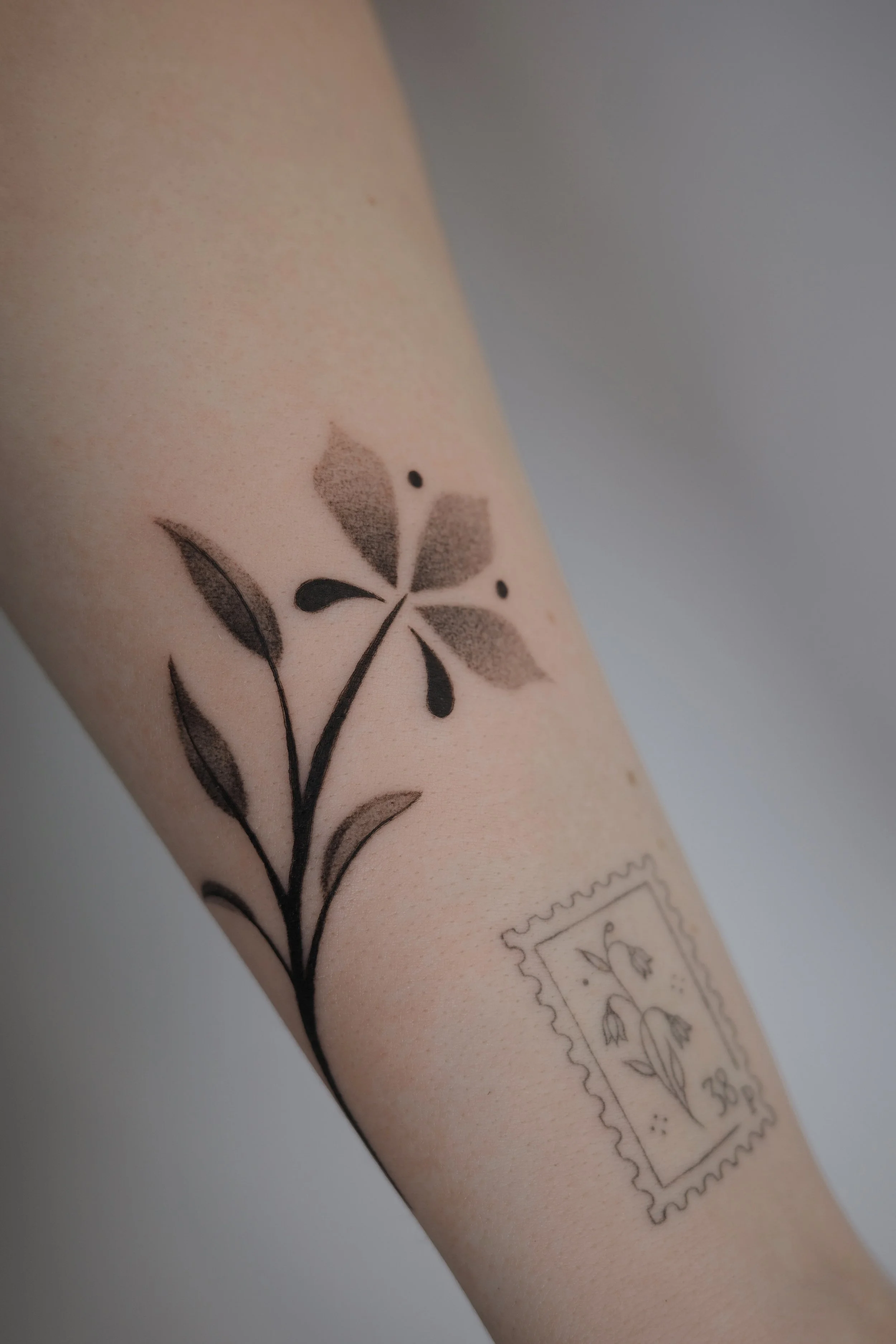 Close-up of a person's arm with a black ink floral tattoo and a postage stamp-style tattoo featuring a flower.