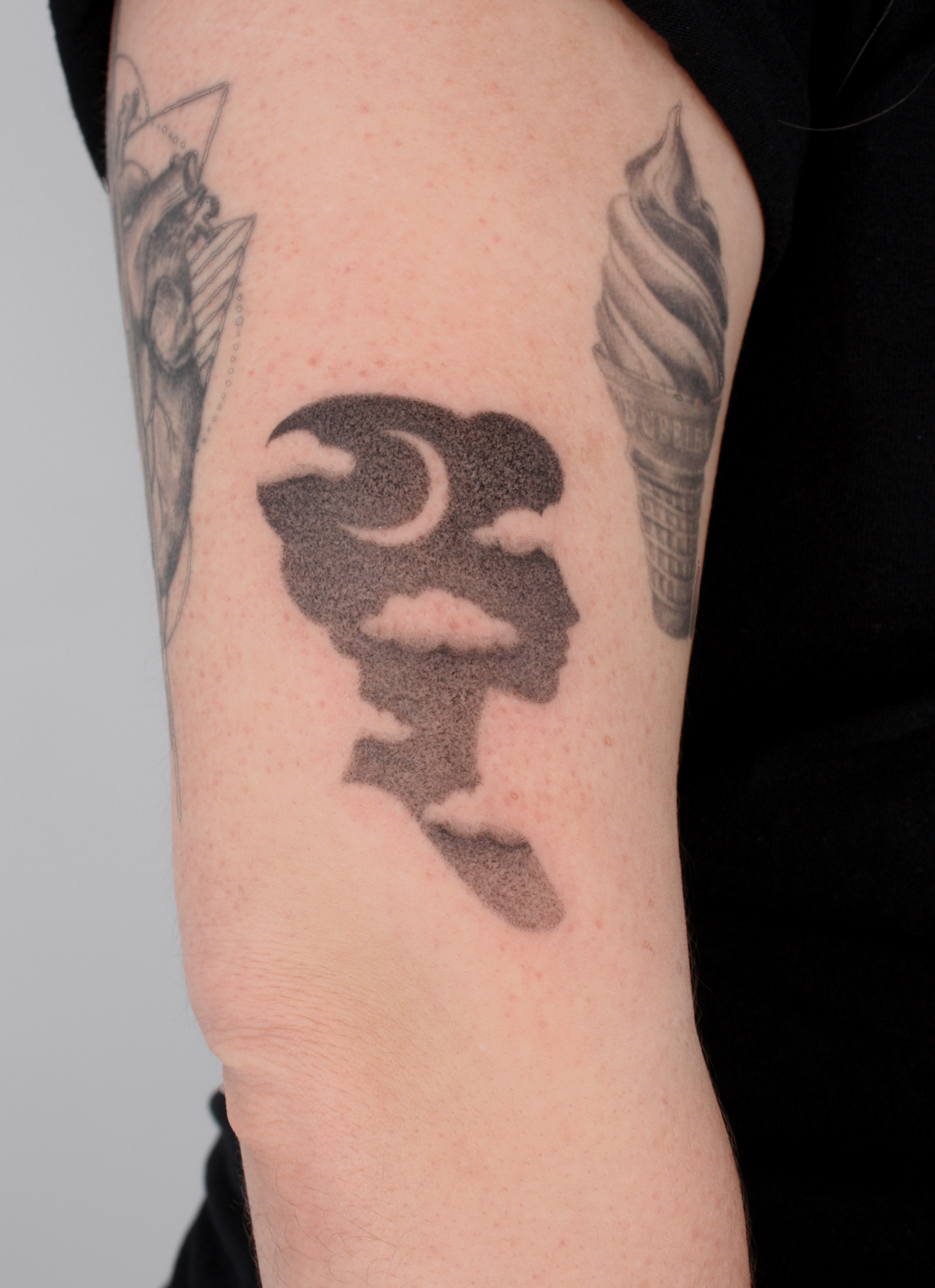 A tattoo on a person's upper arm depicting a stylized face silhouette with thick eyebrows, a mustache, and a beard, rendered in black ink with dot shading.