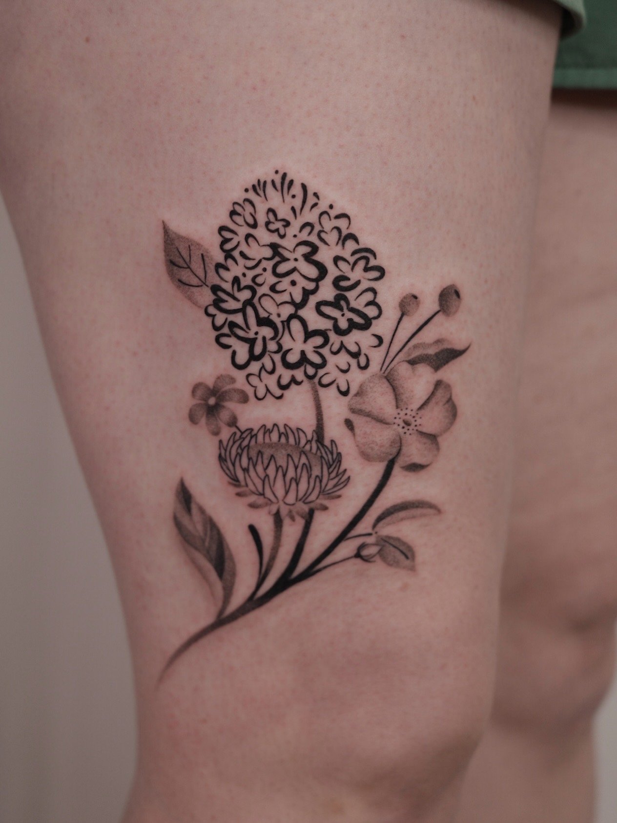 Black and gray floral tattoo on skin featuring a large cluster of small flowers, a dahlia, and a hibiscus with leaves.