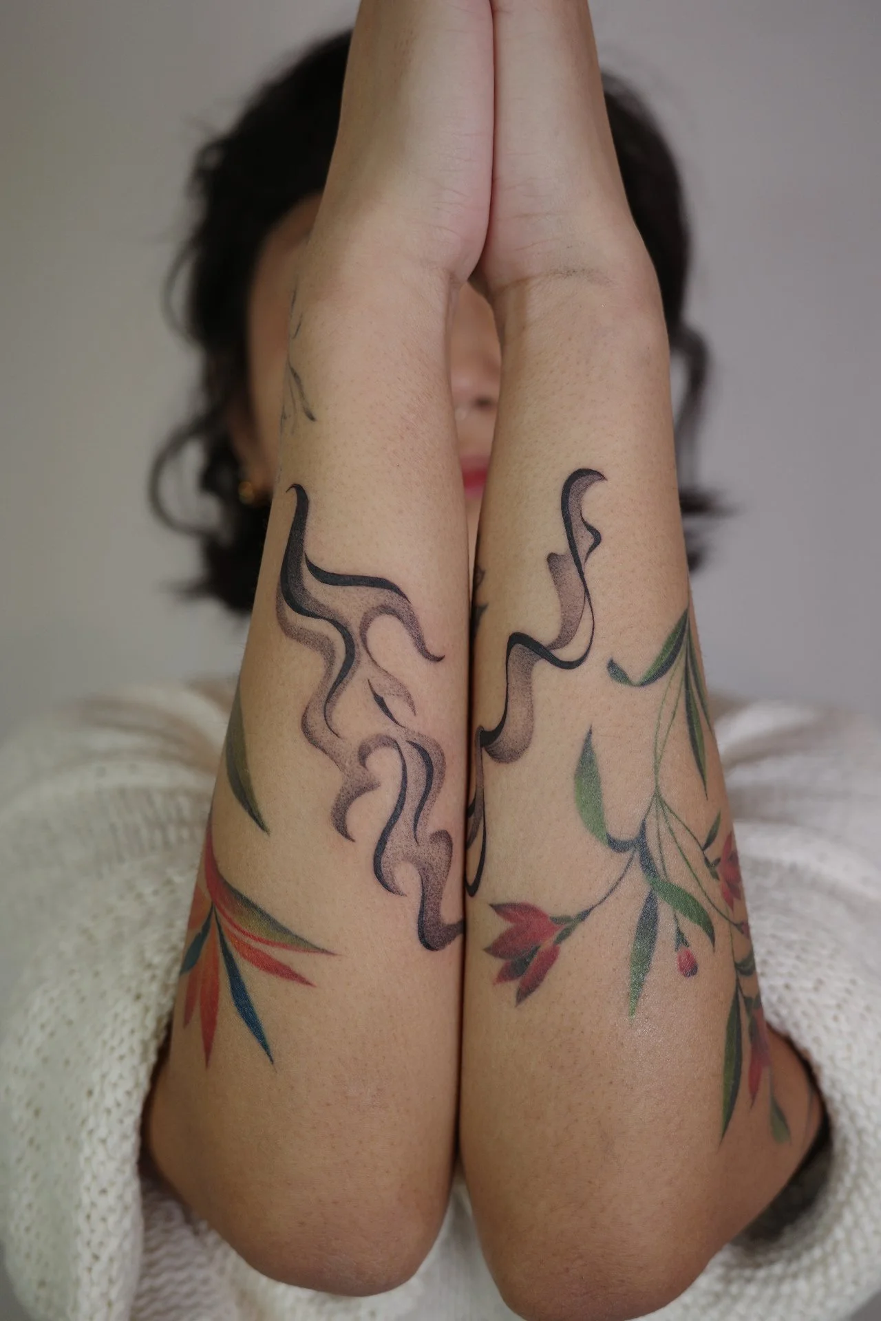 Close-up of person's legs with tattoos and hands pressed together in prayer position, with person's face partially visible in the background.