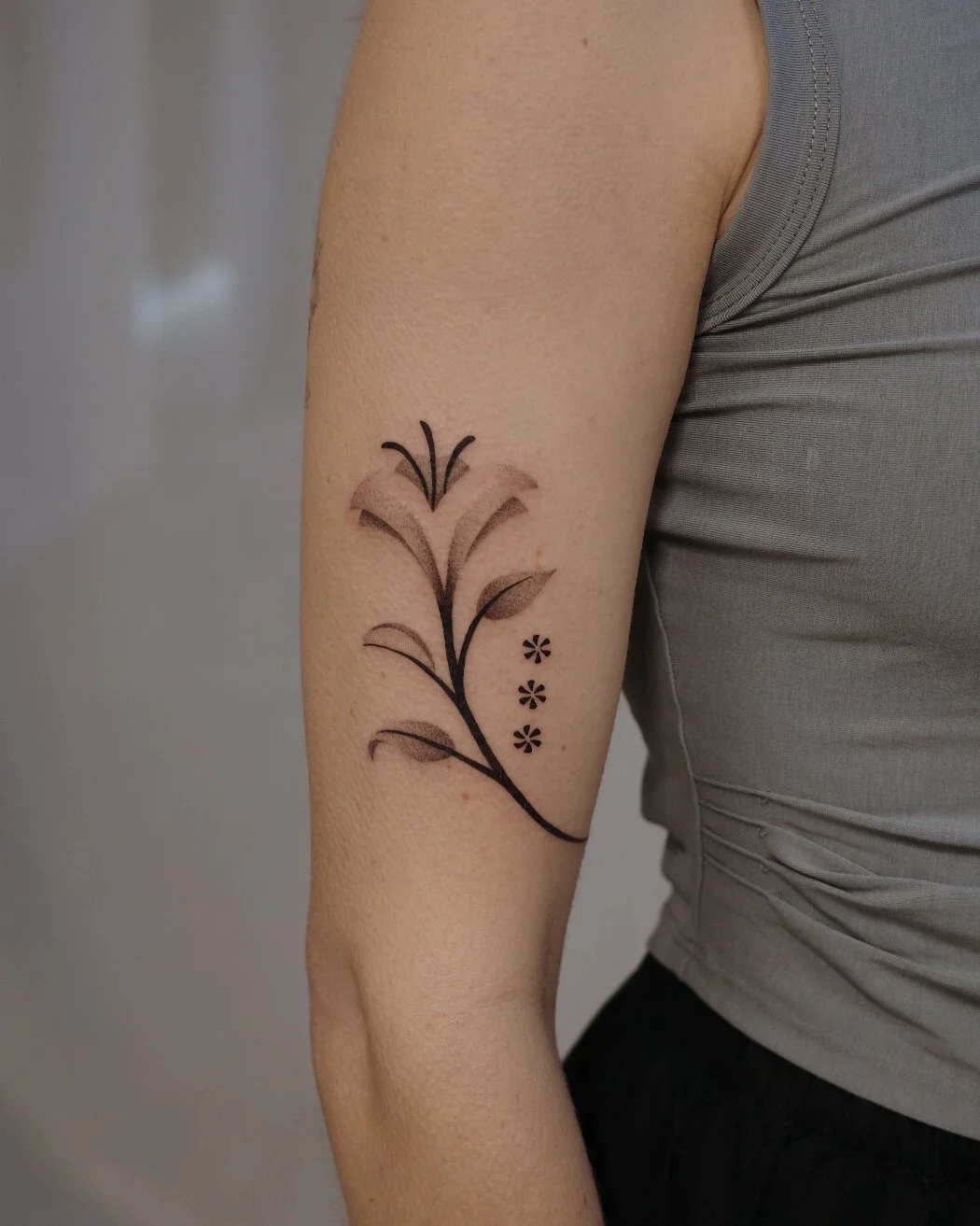 Tattoo of a floral design with black outlines and shading on a person's upper arm, next to a gray sleeveless shirt.