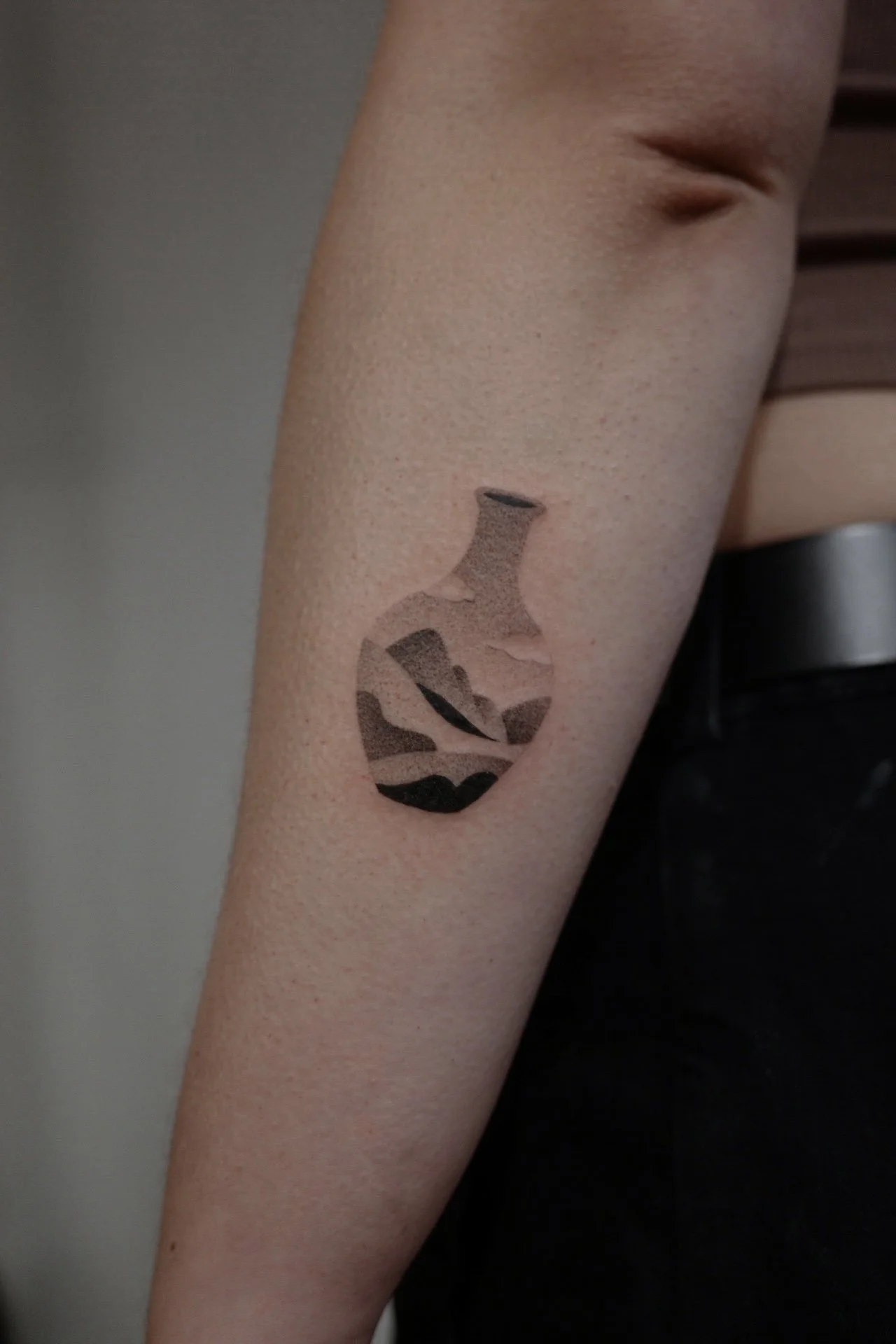 Close-up of a person's arm with a tattoo of a stylized, abstract vase or pitcher in shades of brown, black, and beige.